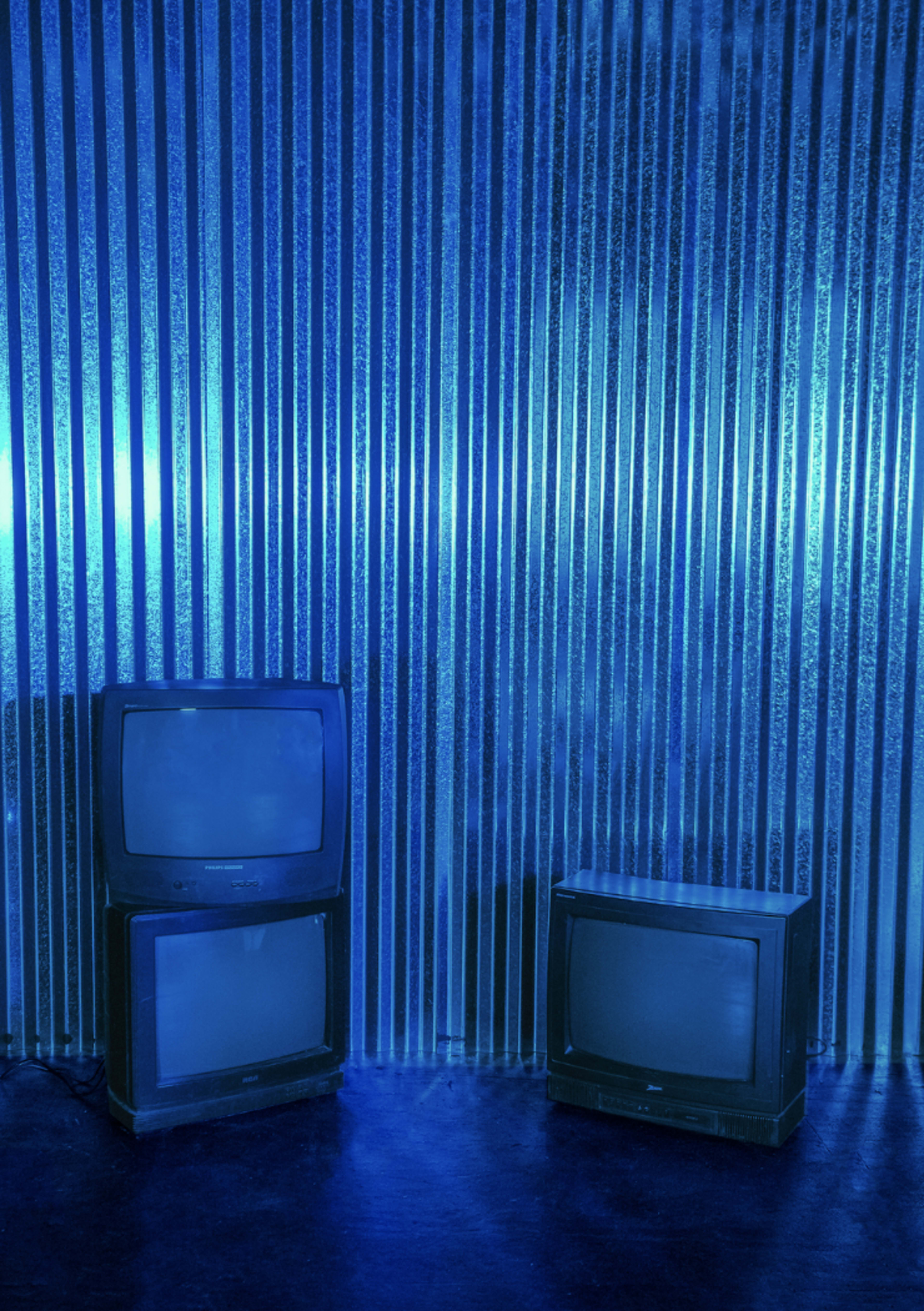 The image shows three retro television sets against a blue corrugated metal wall.