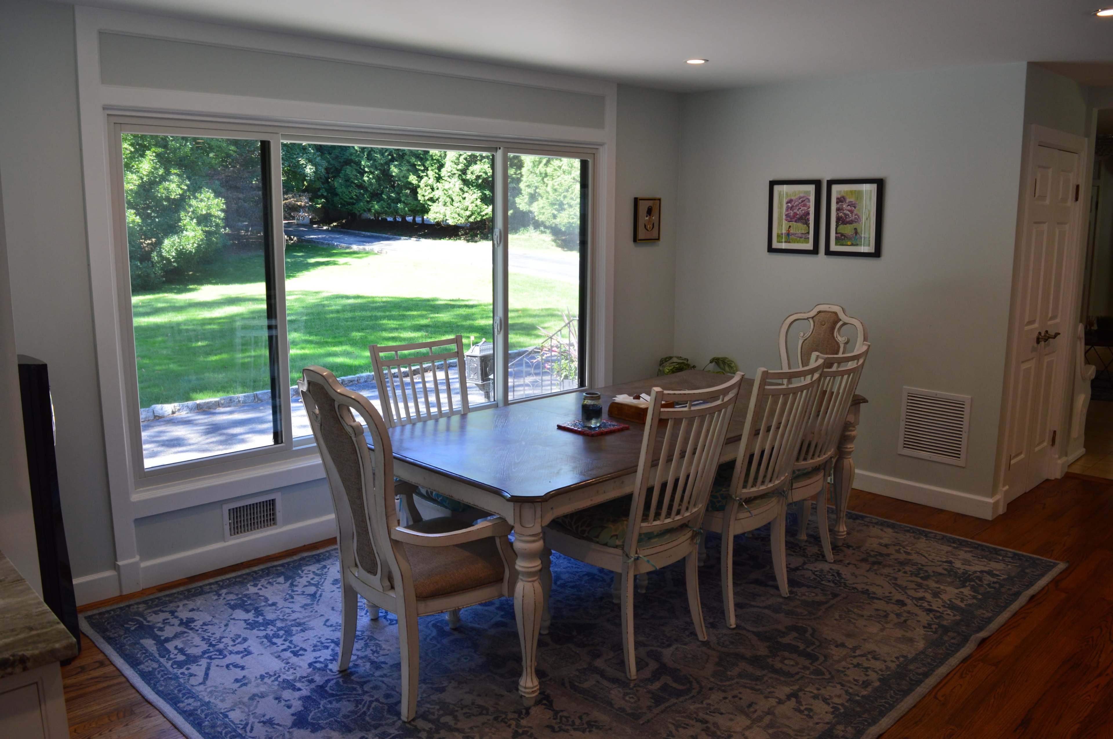A dining room features a rectangular table surrounded by six chairs, positioned near a large window that offers a view of a green lawn and trees outside.