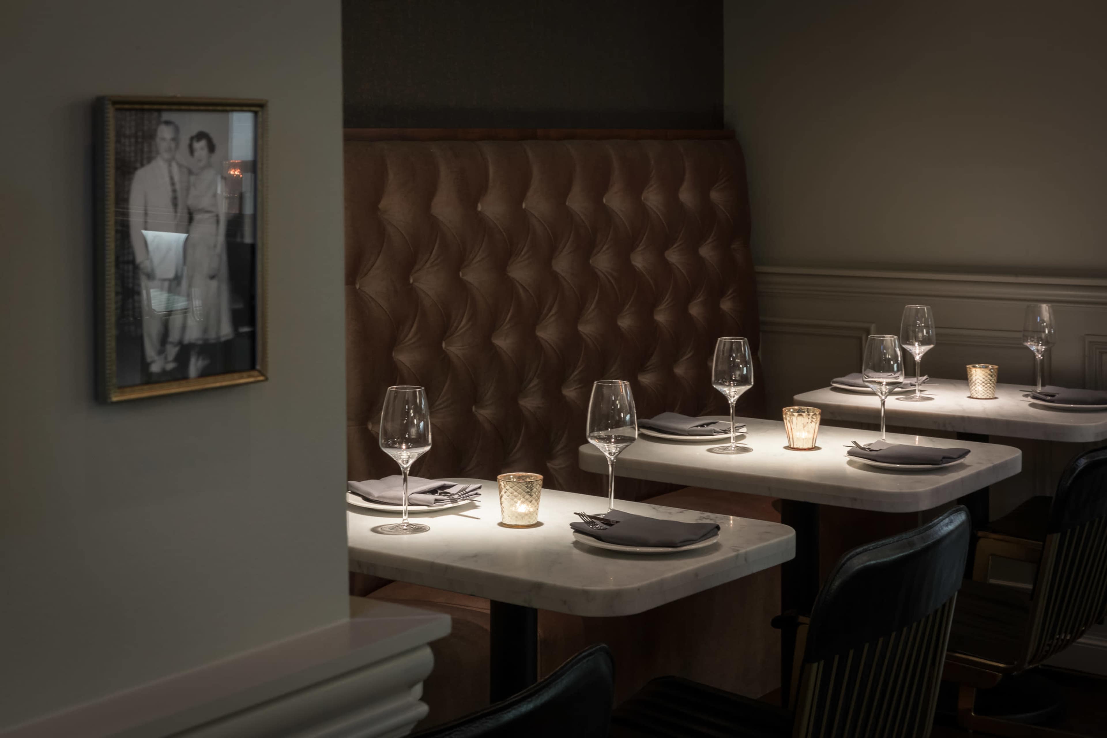 The image features an elegant dining area with a tufted leather banquette, tables set with wine glasses and candles, and a vintage photograph on the wall.