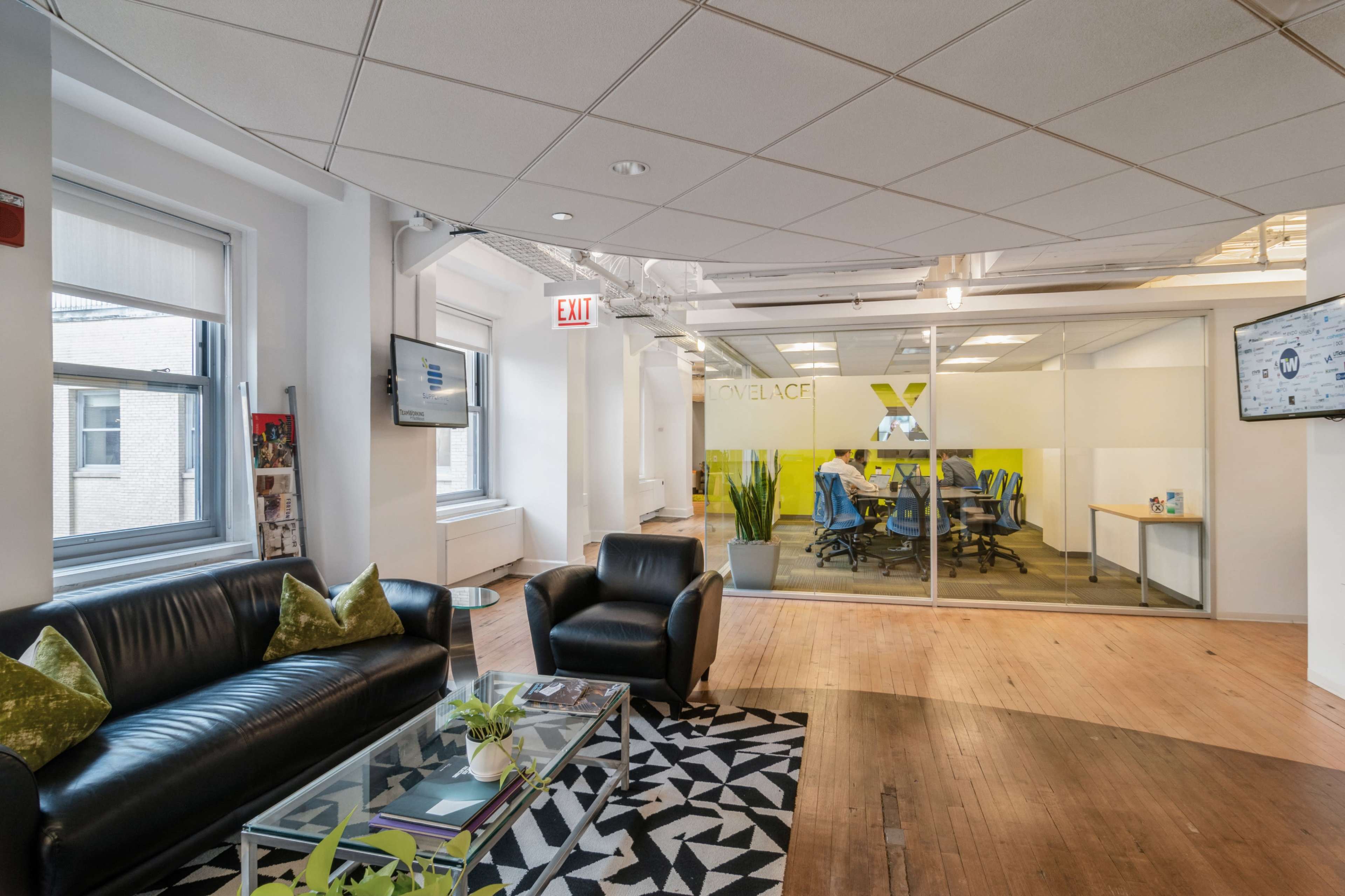 The image shows a modern office space with a black leather sofa and armchair, a glass coffee table, and a conference room visible behind glass walls.