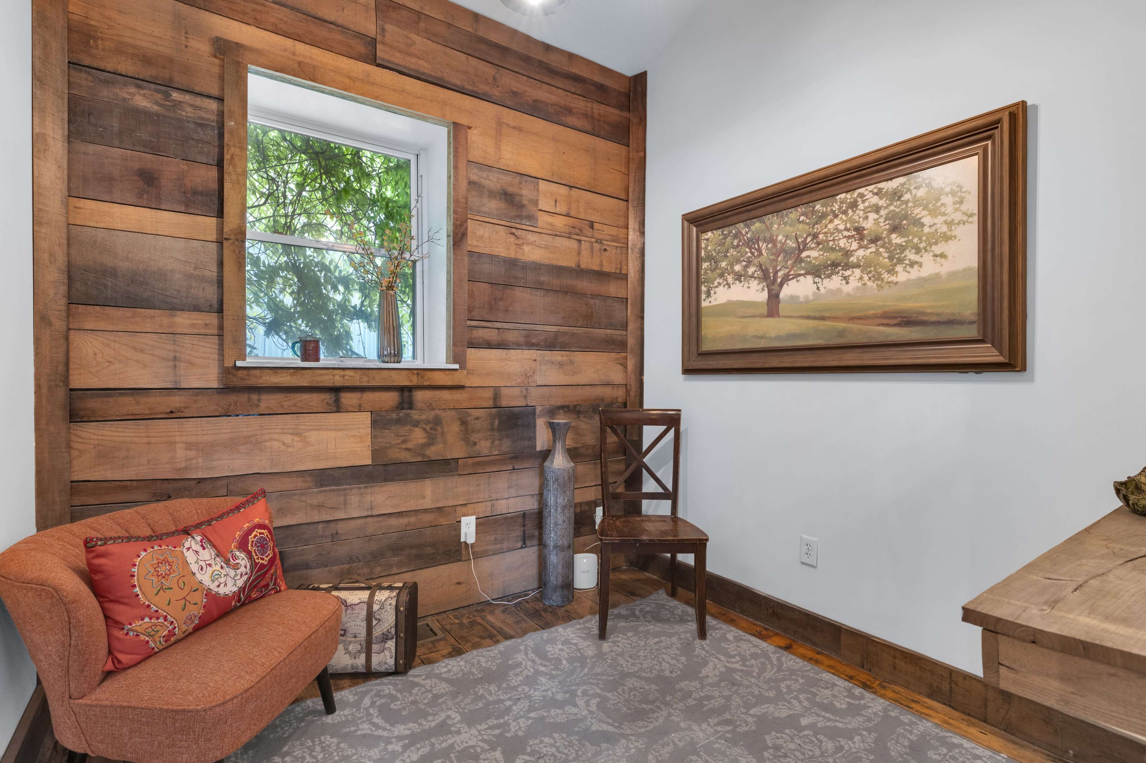 The image shows a cozy corner of a room featuring a wooden accent wall, a small orange armchair with patterned pillows, a framed painting of a tree, and a simple wooden chair.