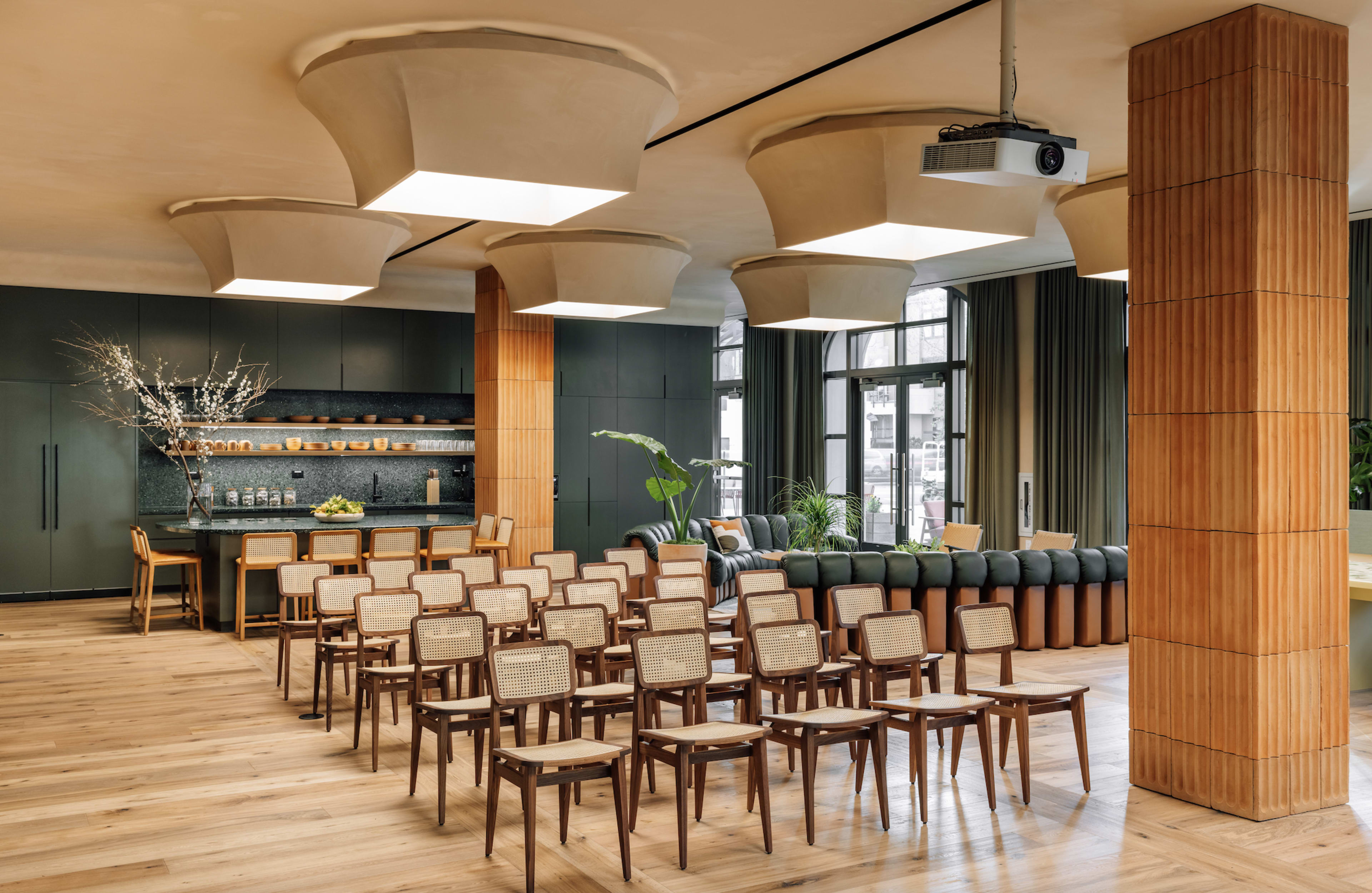 A modern interior space with rows of wooden chairs arranged facing a dark kitchen area, featuring large light fixtures and a mix of seating areas.