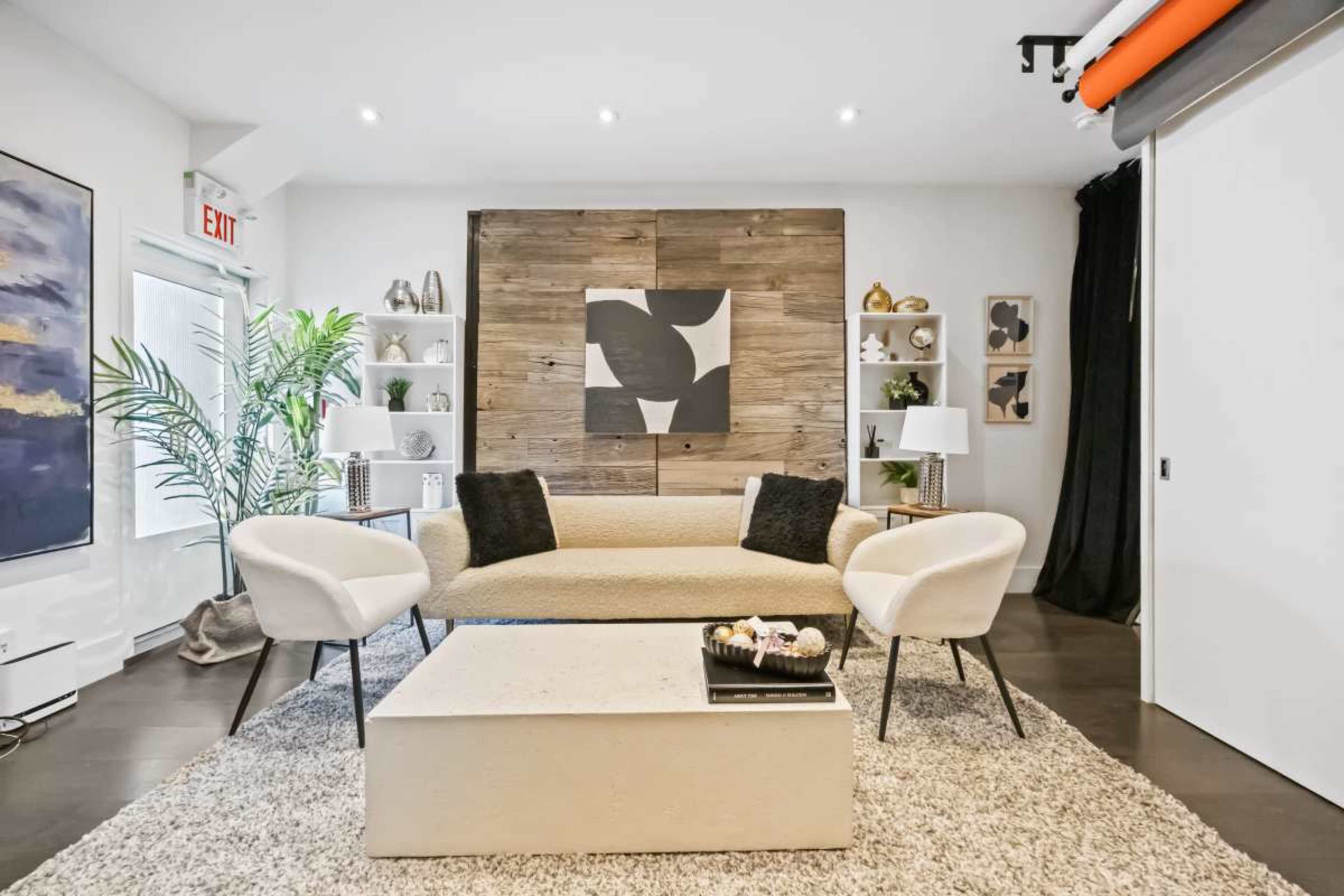 A modern living room features a beige sofa, two light-colored chairs, and a coffee table on a textured rug, with art and decorative items on the walls and shelves.