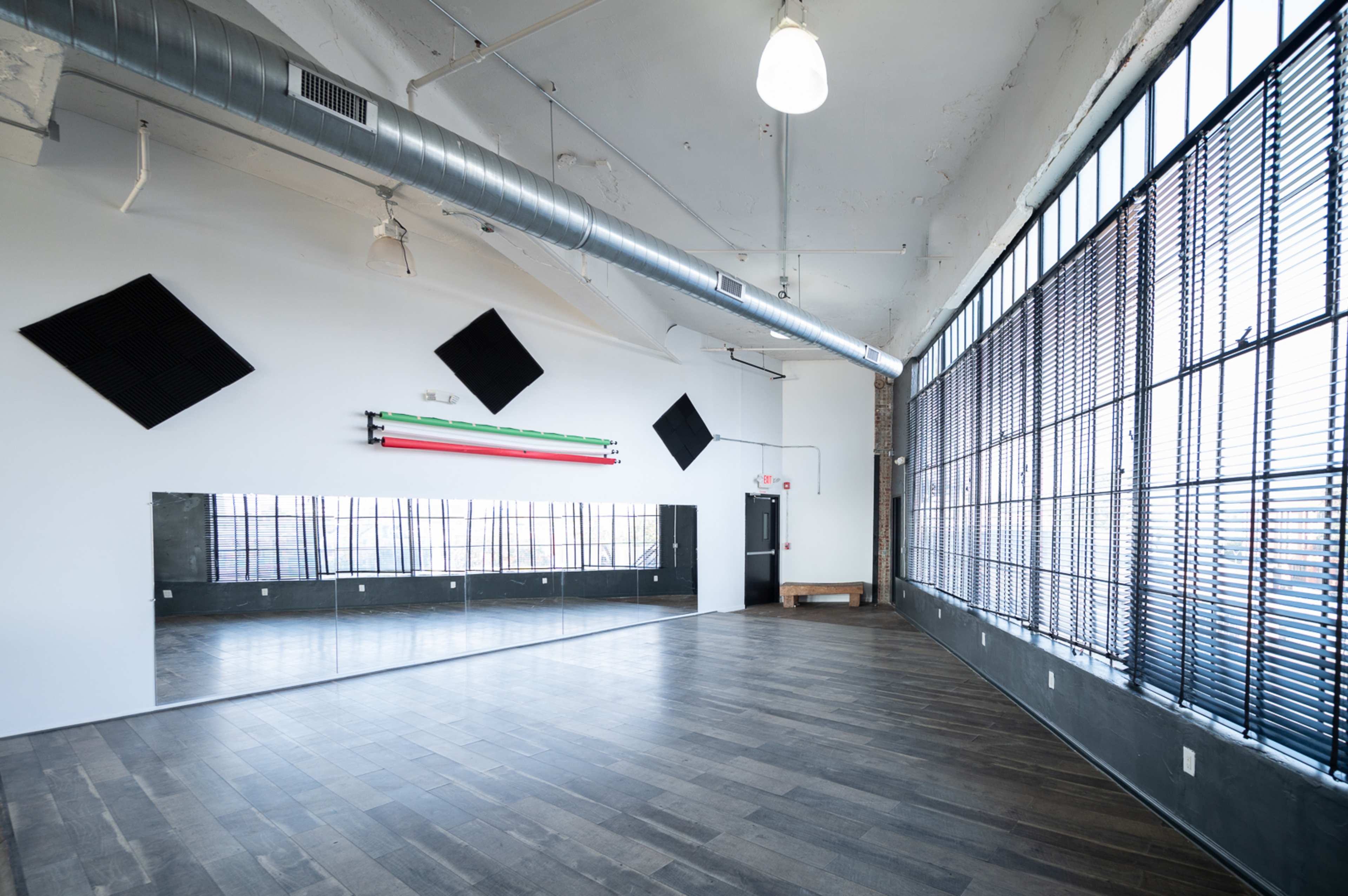 An empty interior space with large windows, a mirrored wall, and wooden flooring.