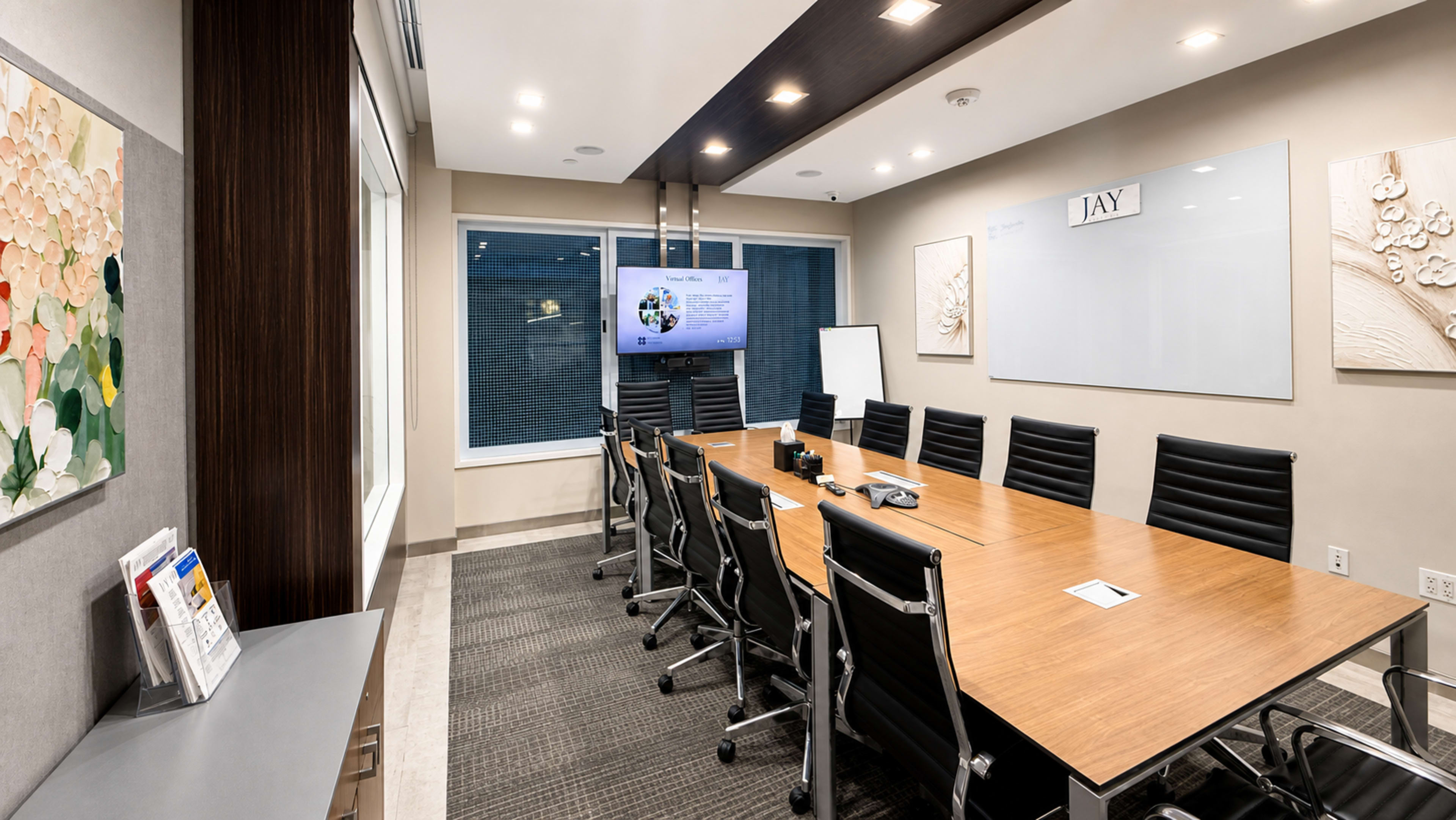 A modern conference room features a long wooden table surrounded by black leather chairs, with a screen displaying a presentation on one wall and large windows providing natural light.