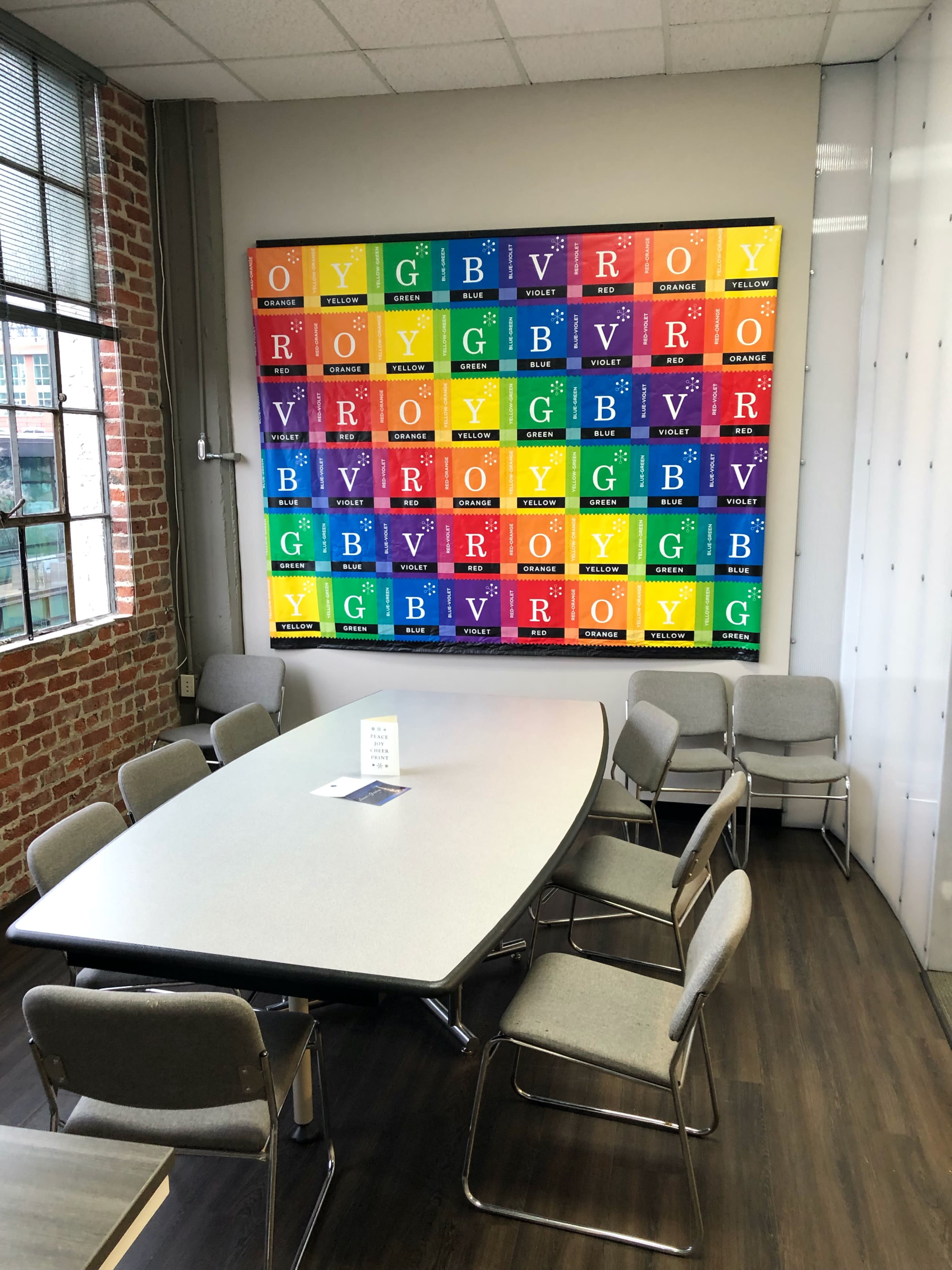 A meeting room with a rectangular table surrounded by chairs, featuring a colorful periodic table artwork mounted on the wall.