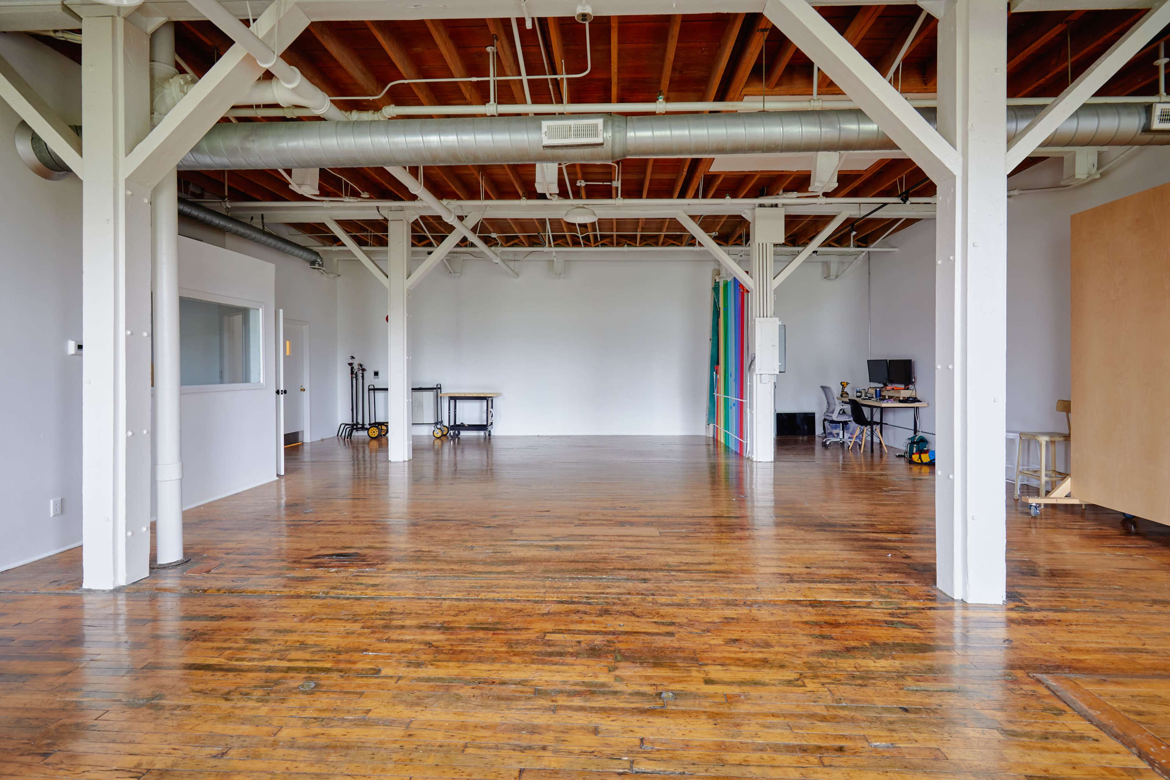 A spacious room with wooden floors, exposed beams, and minimal furnishings, featuring a colorful backdrop on one wall.