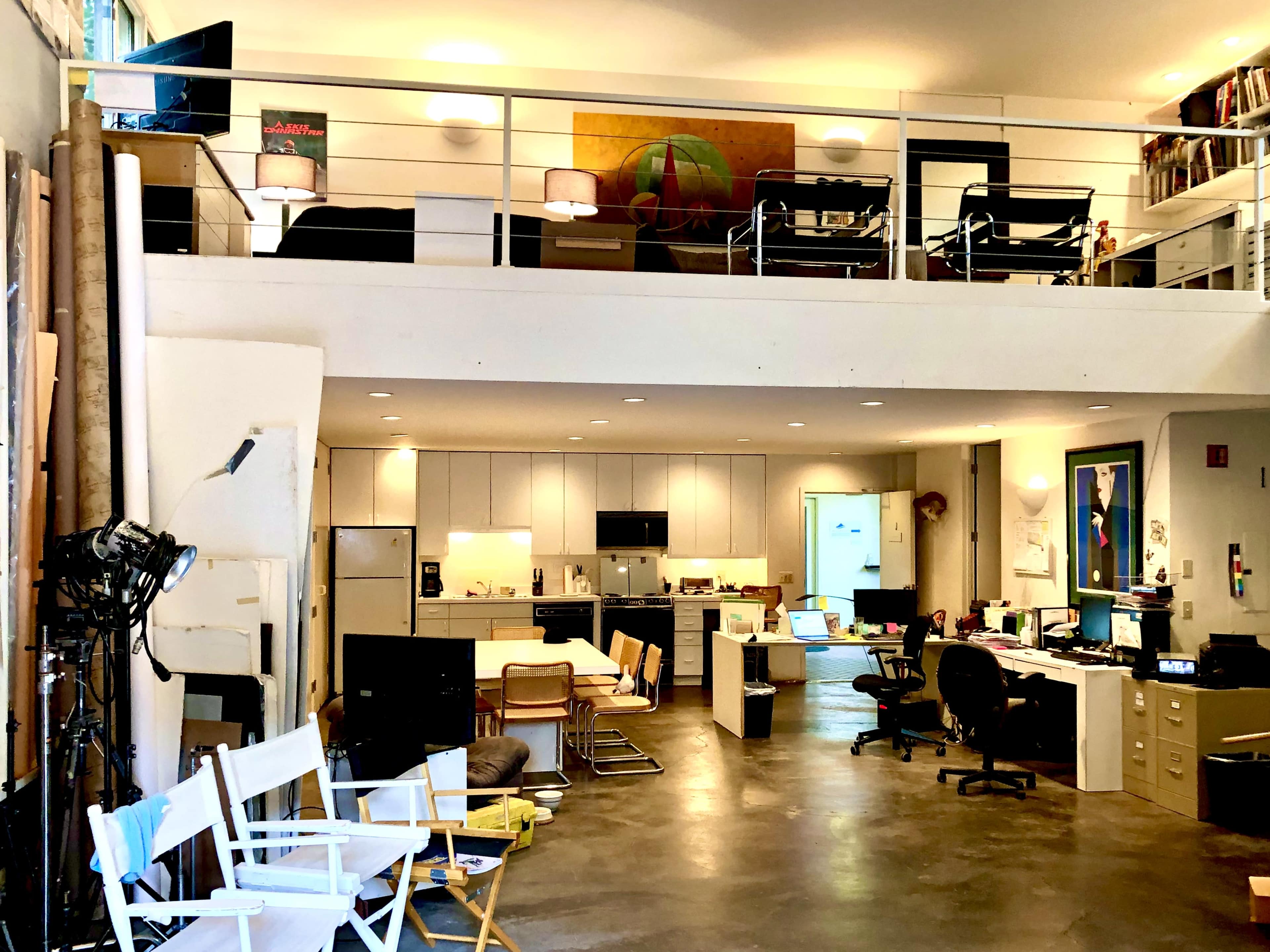 A spacious, multi-level studio apartment featuring a kitchen, workstations, and a loft area with a sitting space.
