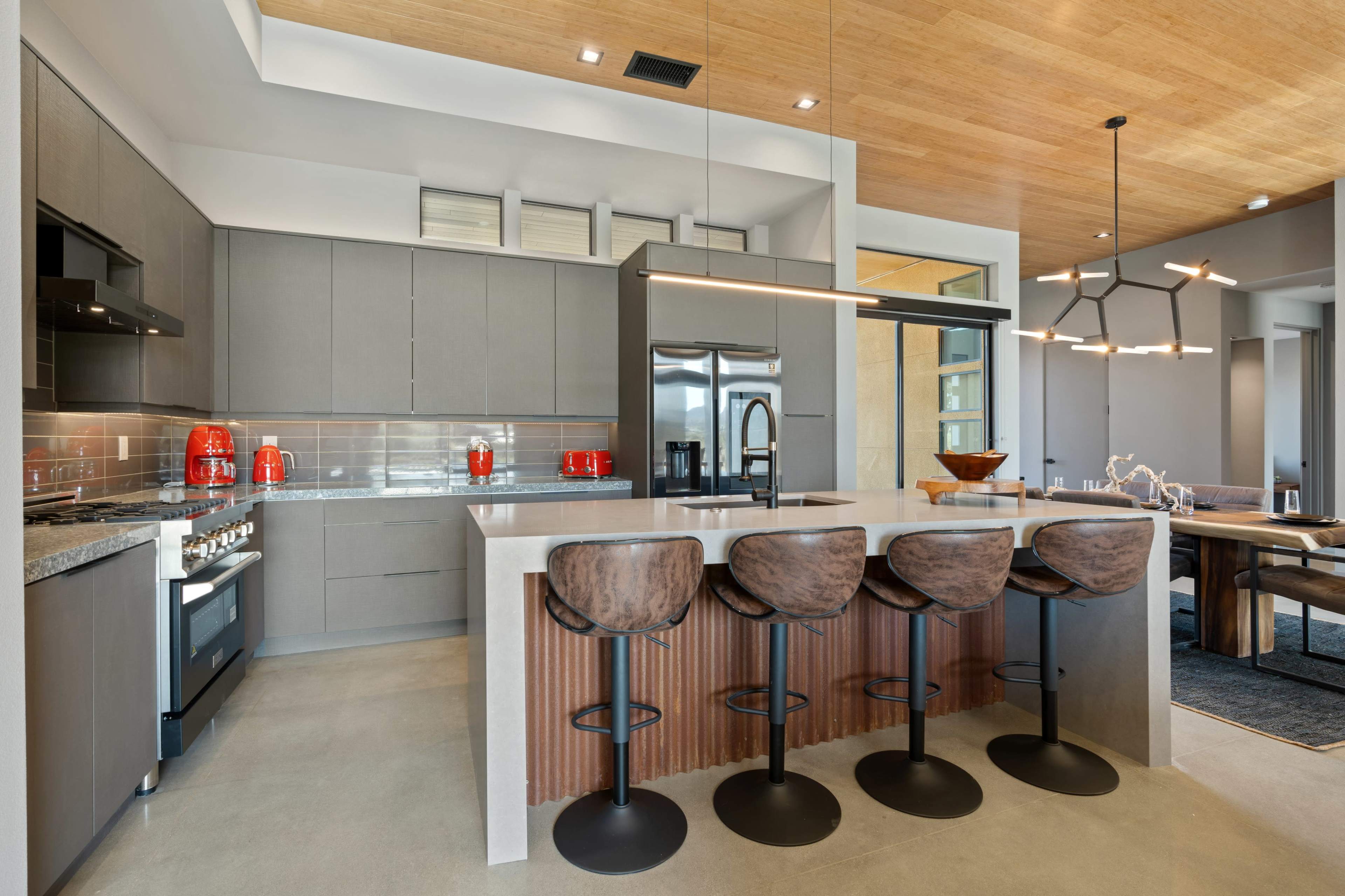 A modern kitchen features a large island with four bar stools, sleek cabinetry, and stainless steel appliances.