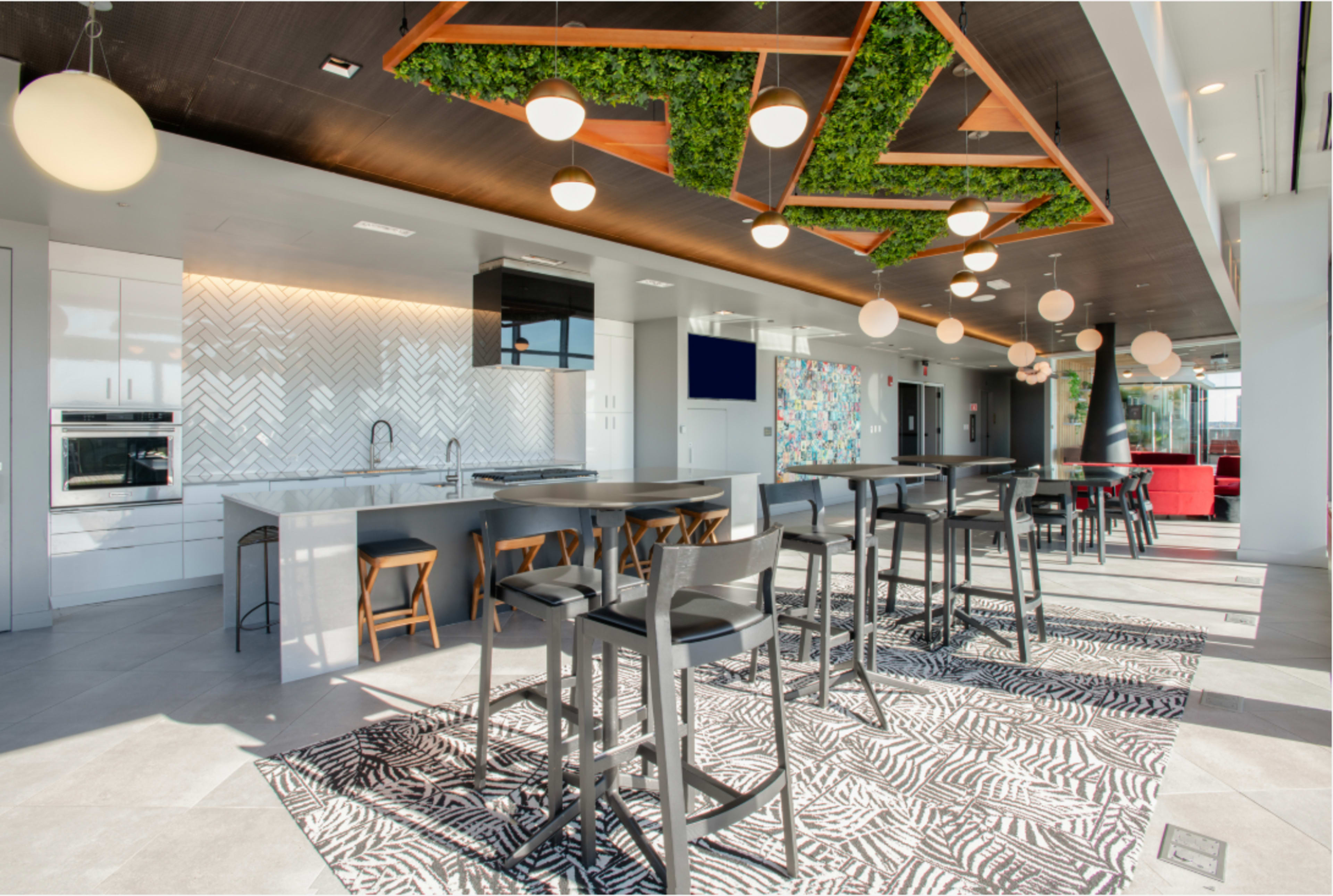 The image shows a modern, open kitchen and dining area with high tables and stools, featuring a geometric ceiling design with greenery and large windows allowing natural light.