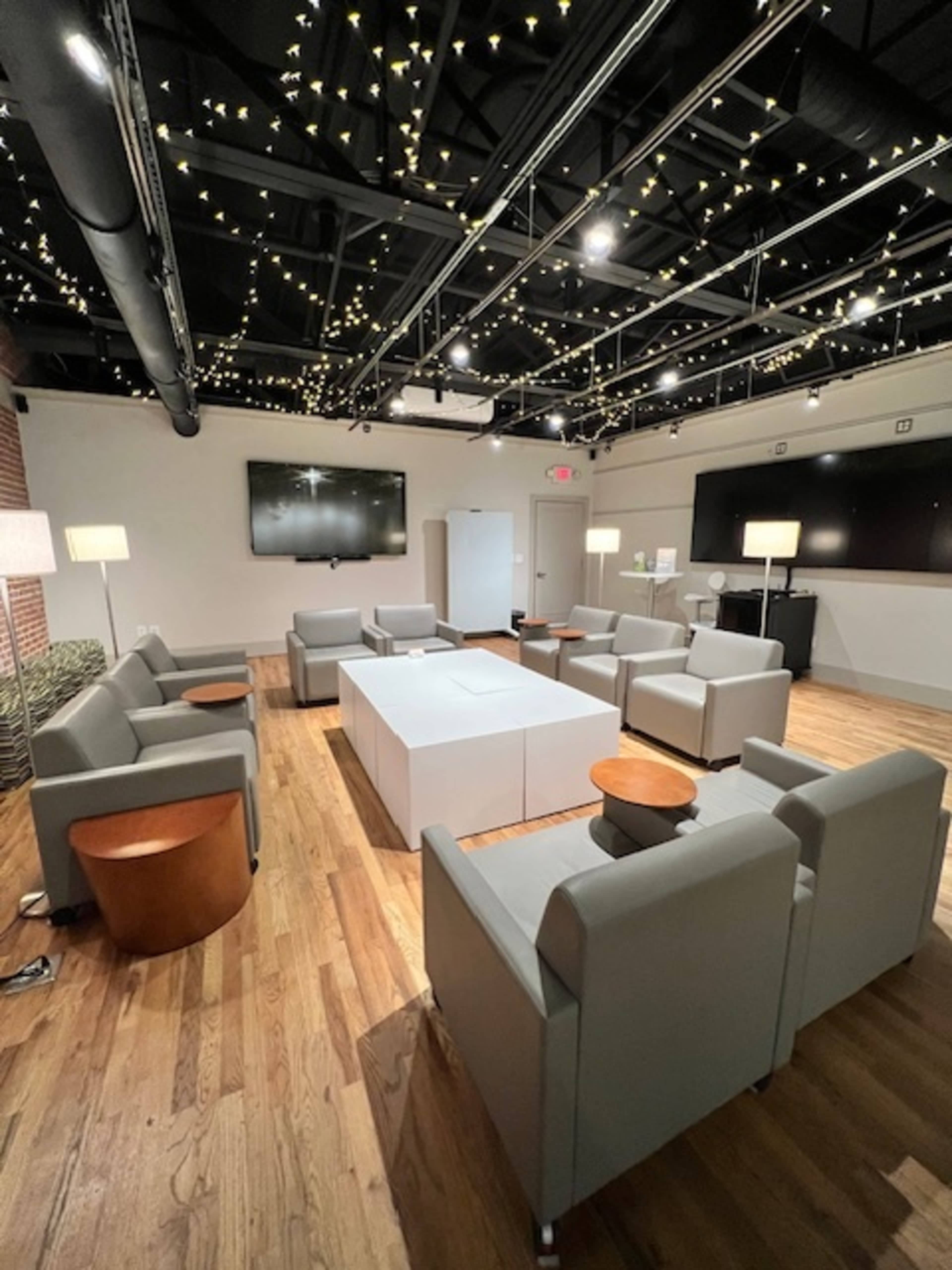 The image shows a spacious lounge area with gray seating arranged around a central white table, illuminated by string lights on the ceiling.