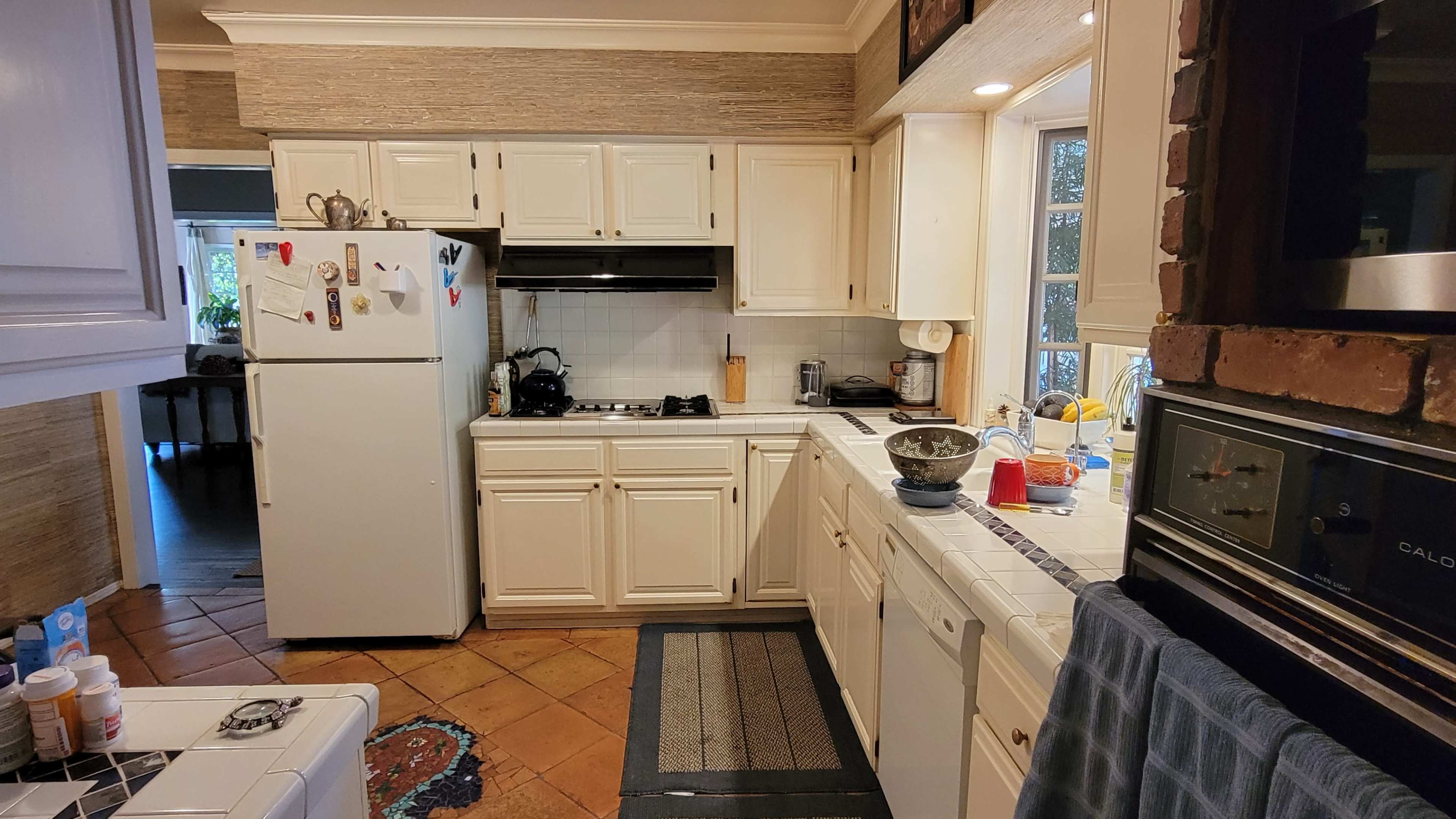 A kitchen features white cabinets, a black stove, a refrigerator covered with magnets and notes, and a tiled countertop with cooking supplies.