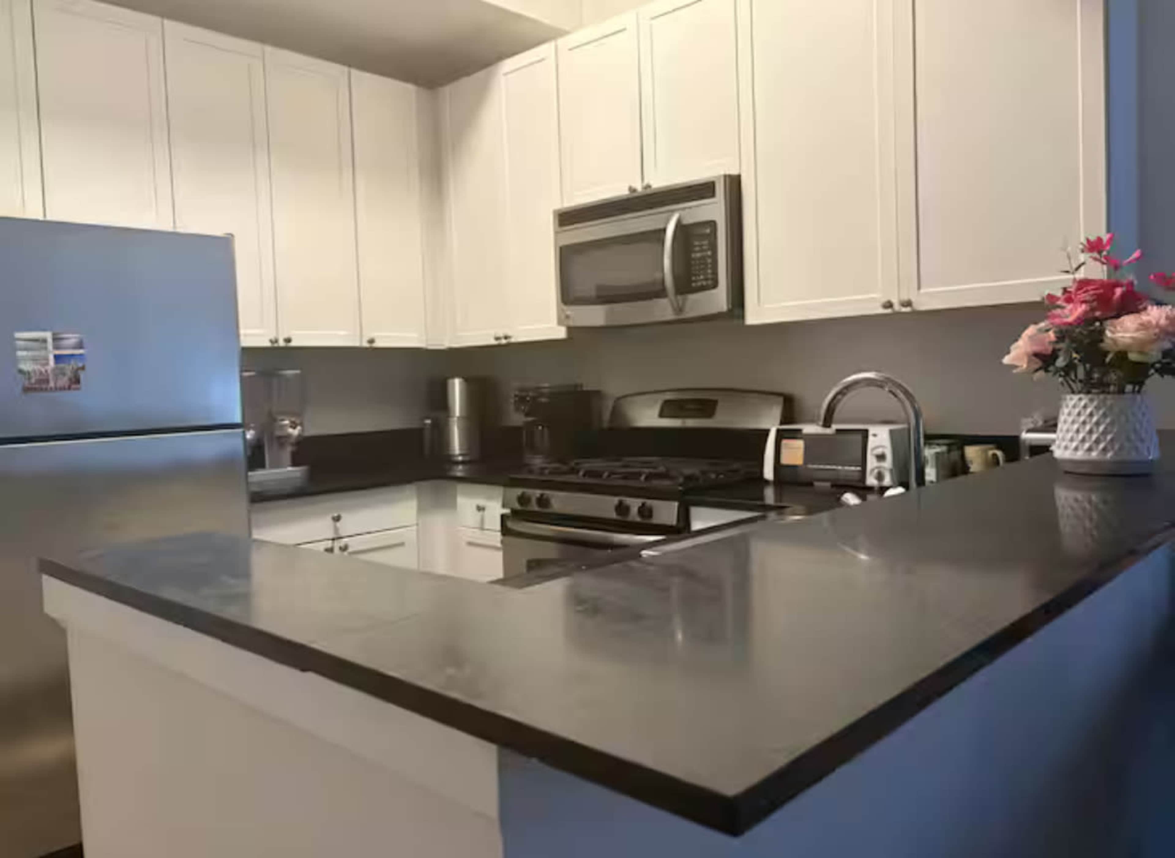 The image shows a modern kitchen with white cabinets, a stainless steel refrigerator, and a black countertop featuring a gas stove and microwave.