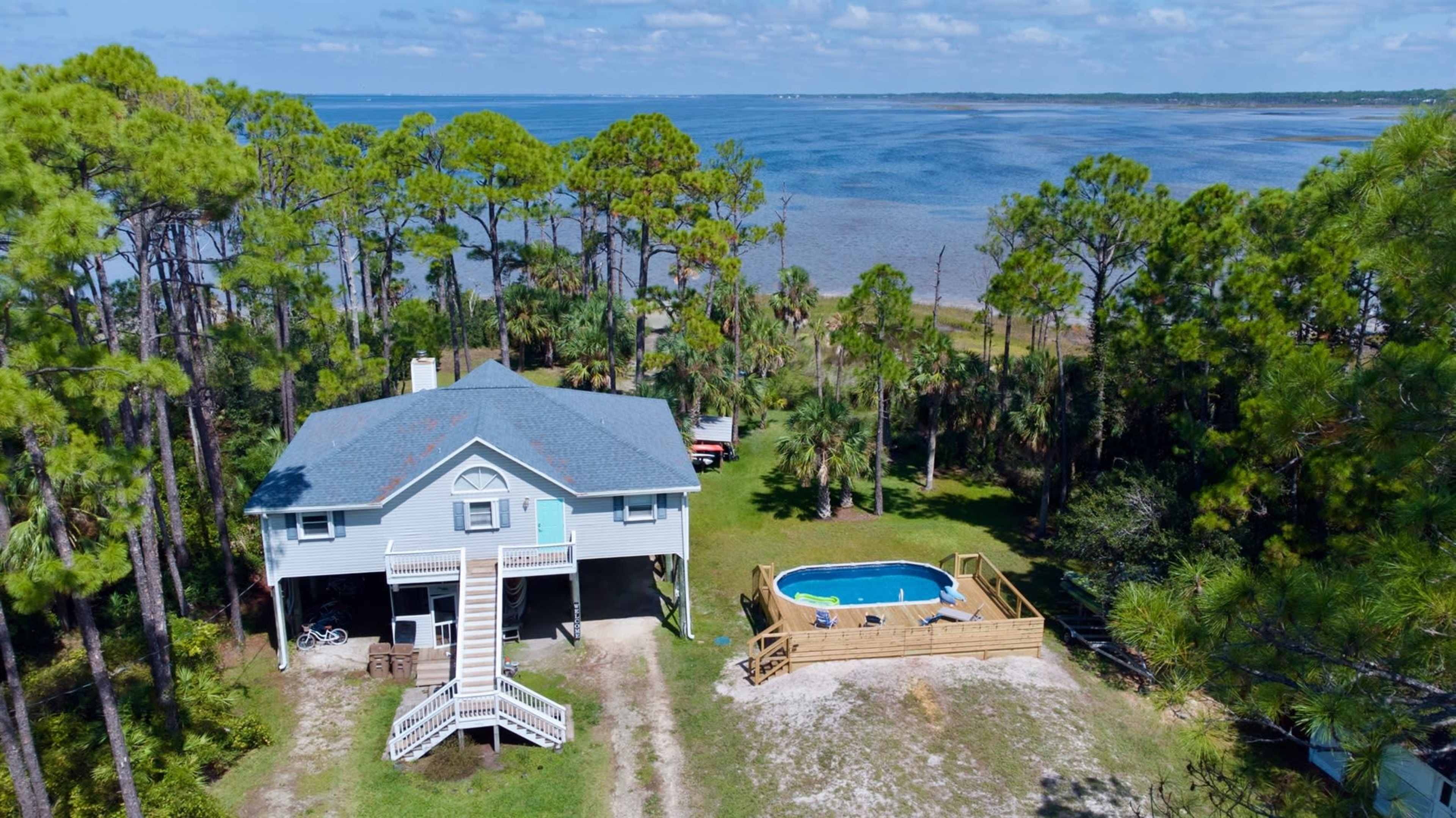 A two-story house with a gray roof is situated near a body of water, surrounded by tall pine trees and featuring a small above-ground pool in the yard.