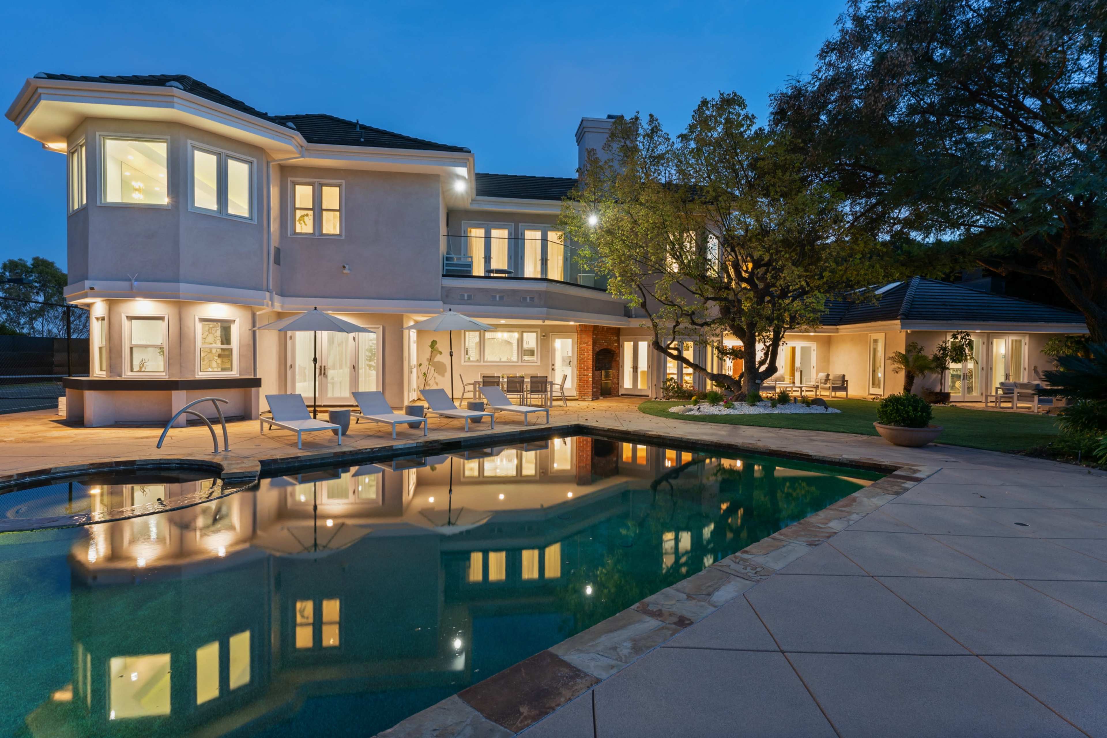 A large, modern two-story house with a swimming pool and lounge chairs is illuminated at dusk, reflecting in the water.