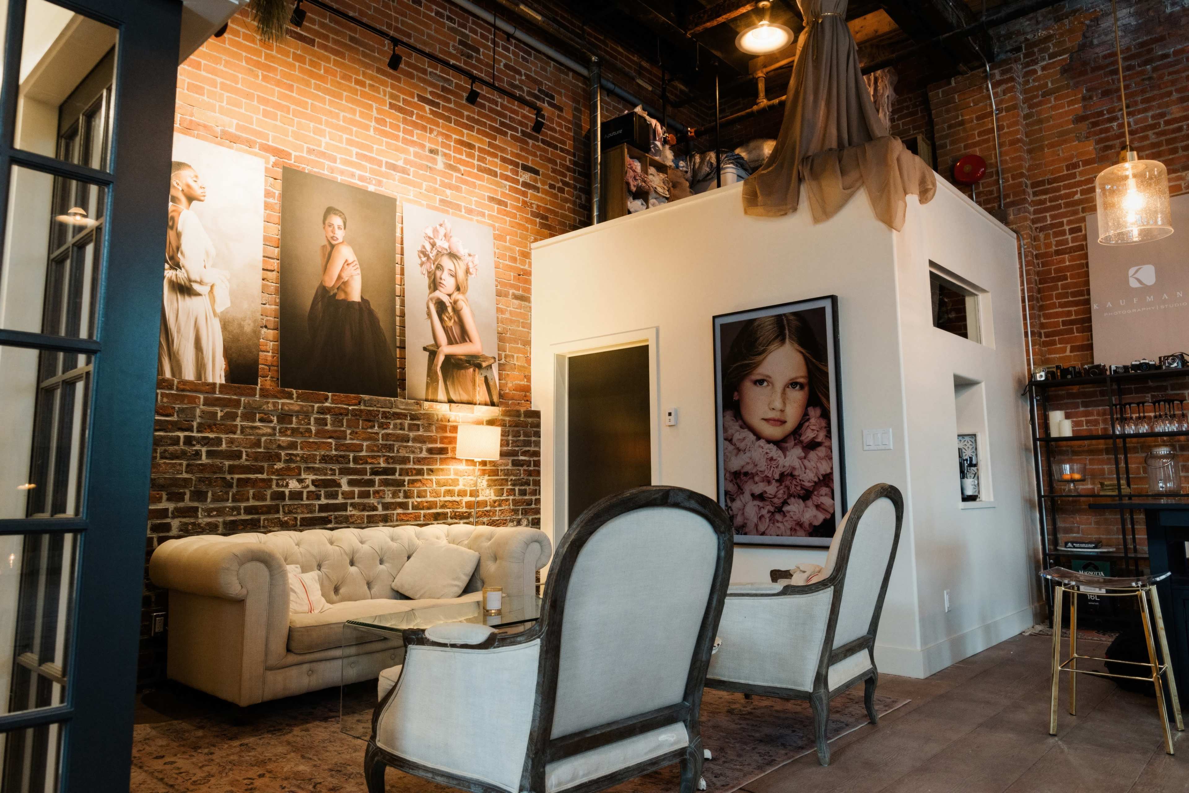 The scene features a modern interior with exposed brick walls, a tufted beige sofa, and several framed portraits of women displayed on the walls.