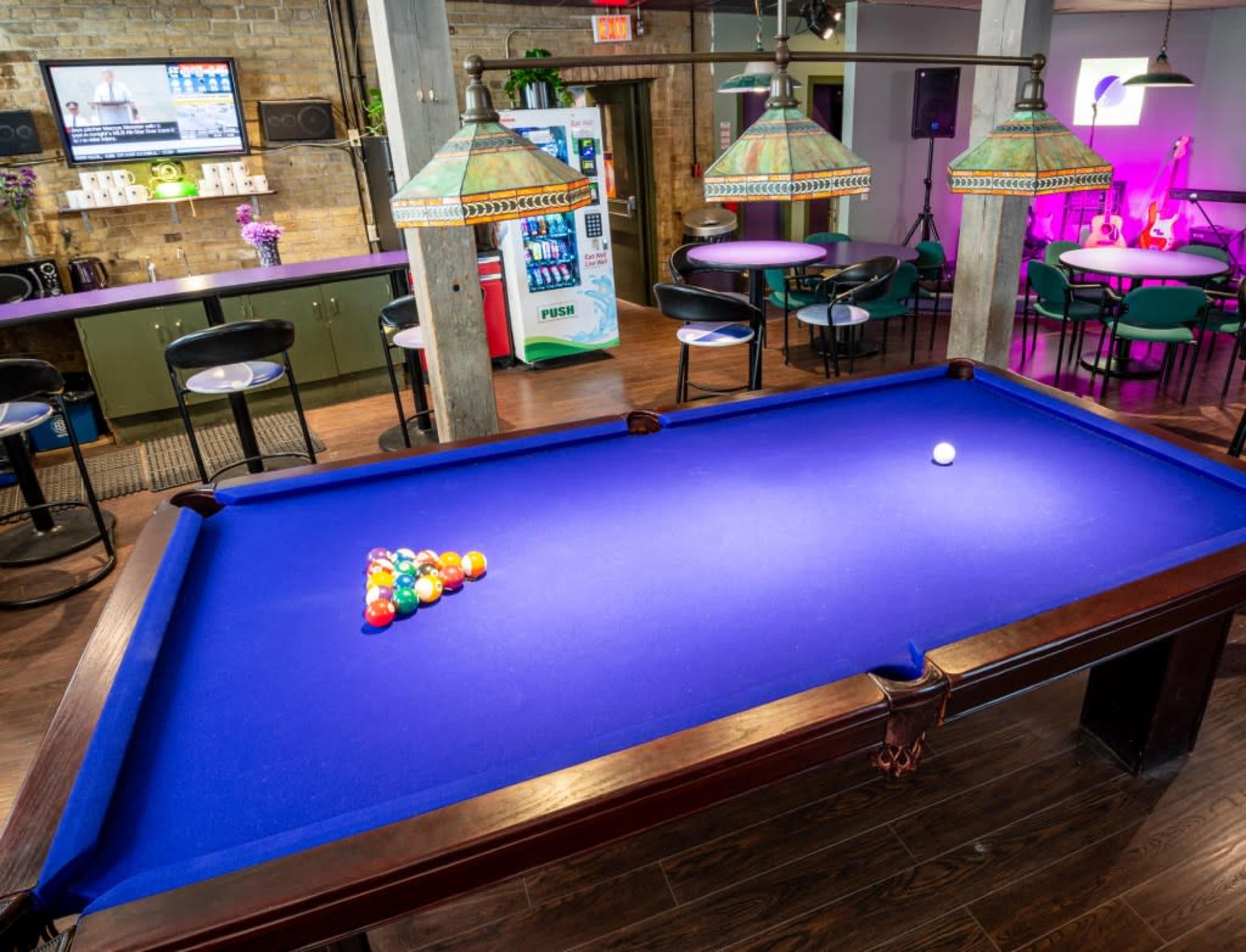 A billiards table with a triangle of colored balls set on its blue felt surface is positioned in a modern recreational room with pendant lighting and lounge seating.