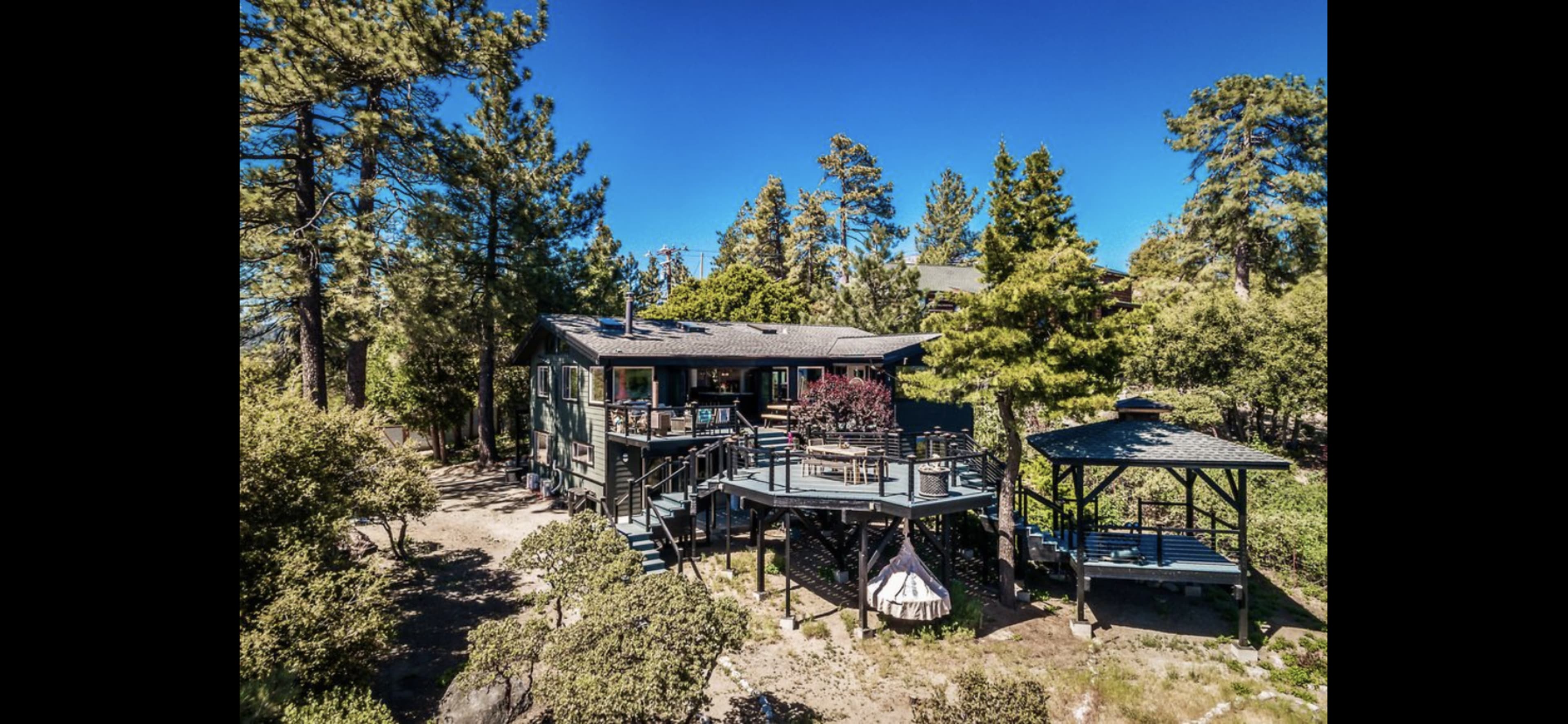A modern two-story house with a wraparound deck and a gazebo is situated among tall trees in a wooded area.