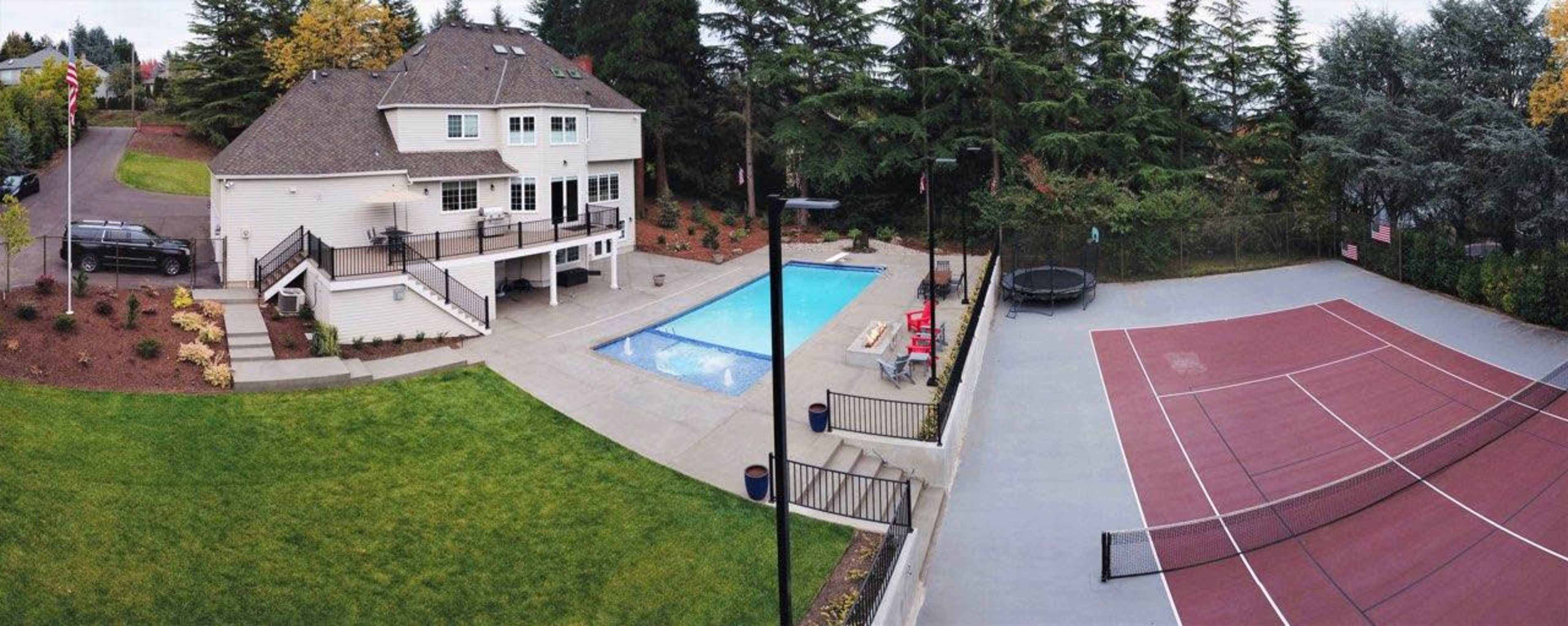 The image shows a large two-story house with a swimming pool, a trampoline, and a tennis court in a landscaped yard surrounded by trees.