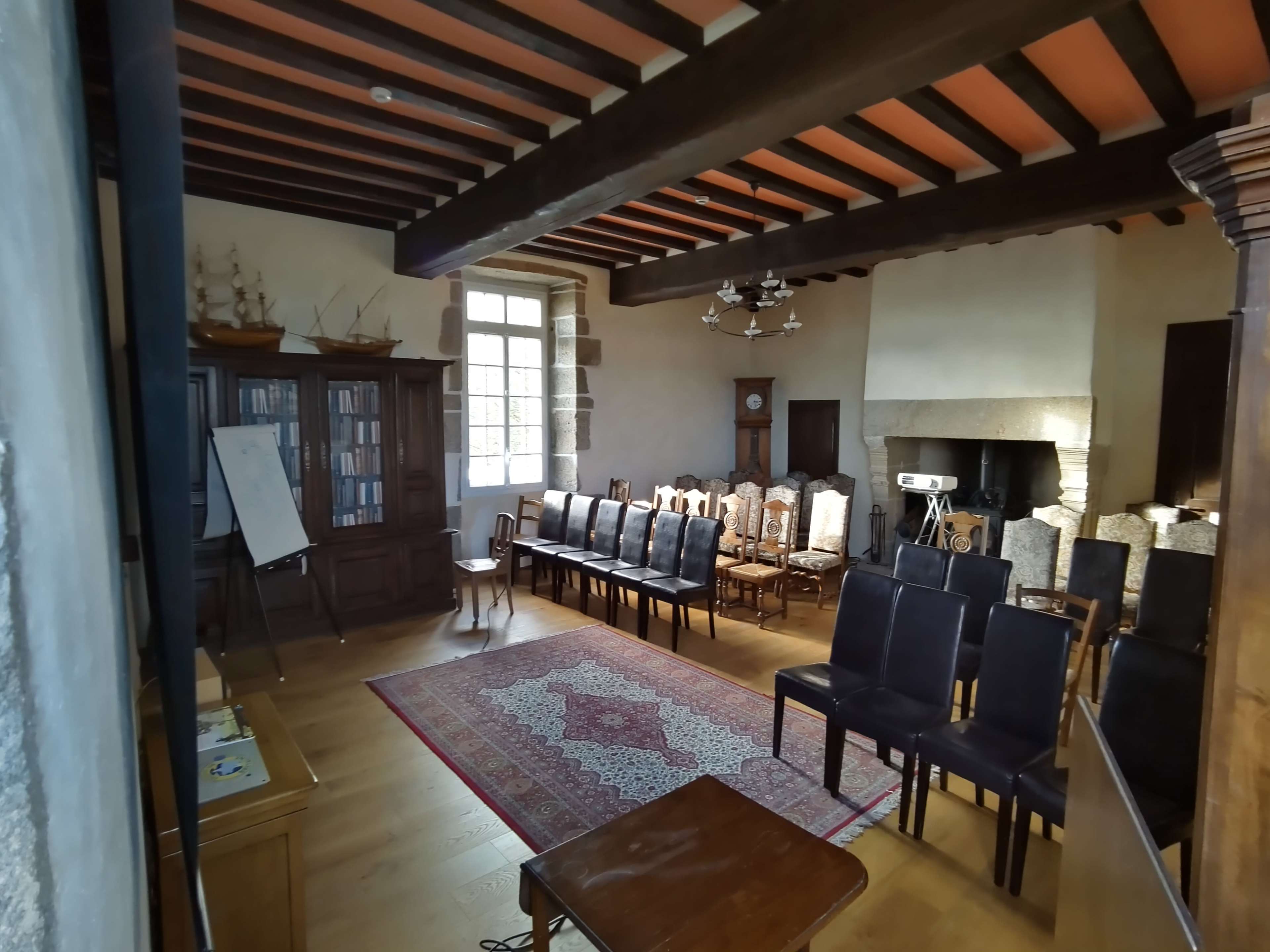 The image shows a spacious room furnished with rows of black chairs, a large wooden bookcase, and decorative elements such as a rug and wall-mounted lighting fixtures.