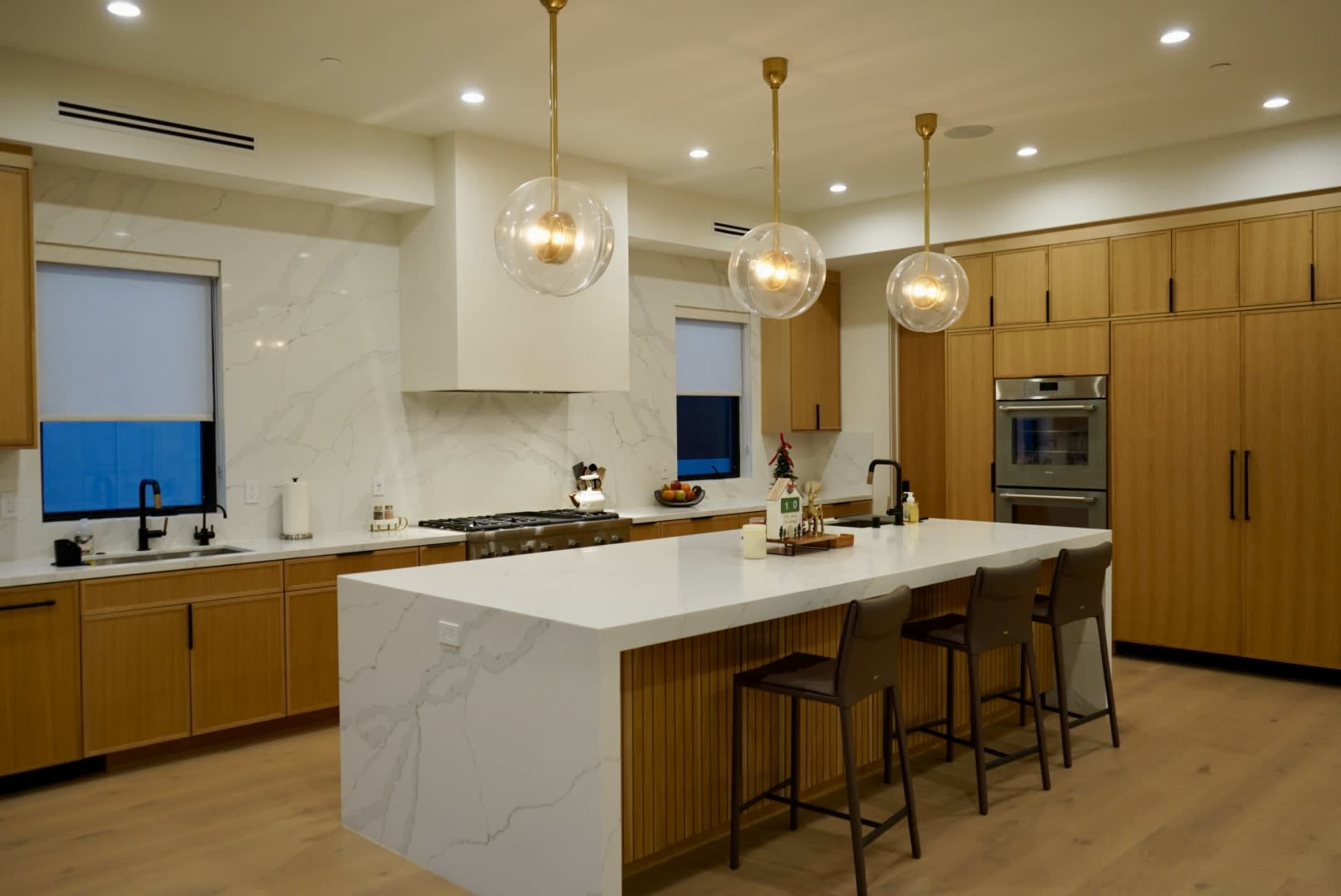 The image shows a modern kitchen with light wood cabinetry, a large white marble countertop, and three pendant lights hanging above a breakfast bar with four chairs.