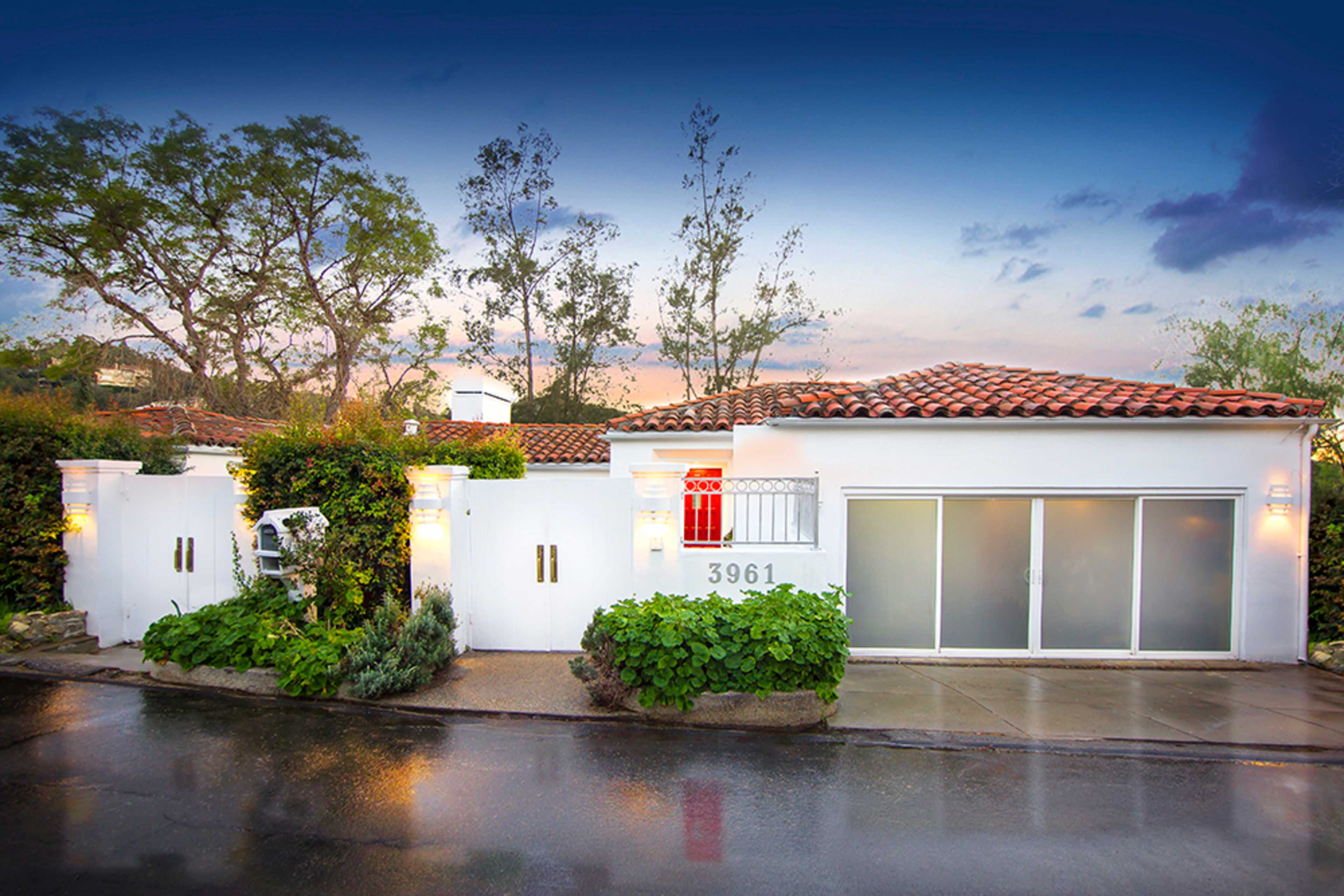 A white house with a tiled roof, situated behind a low wall and flanked by greenery, on a street with wet pavement.