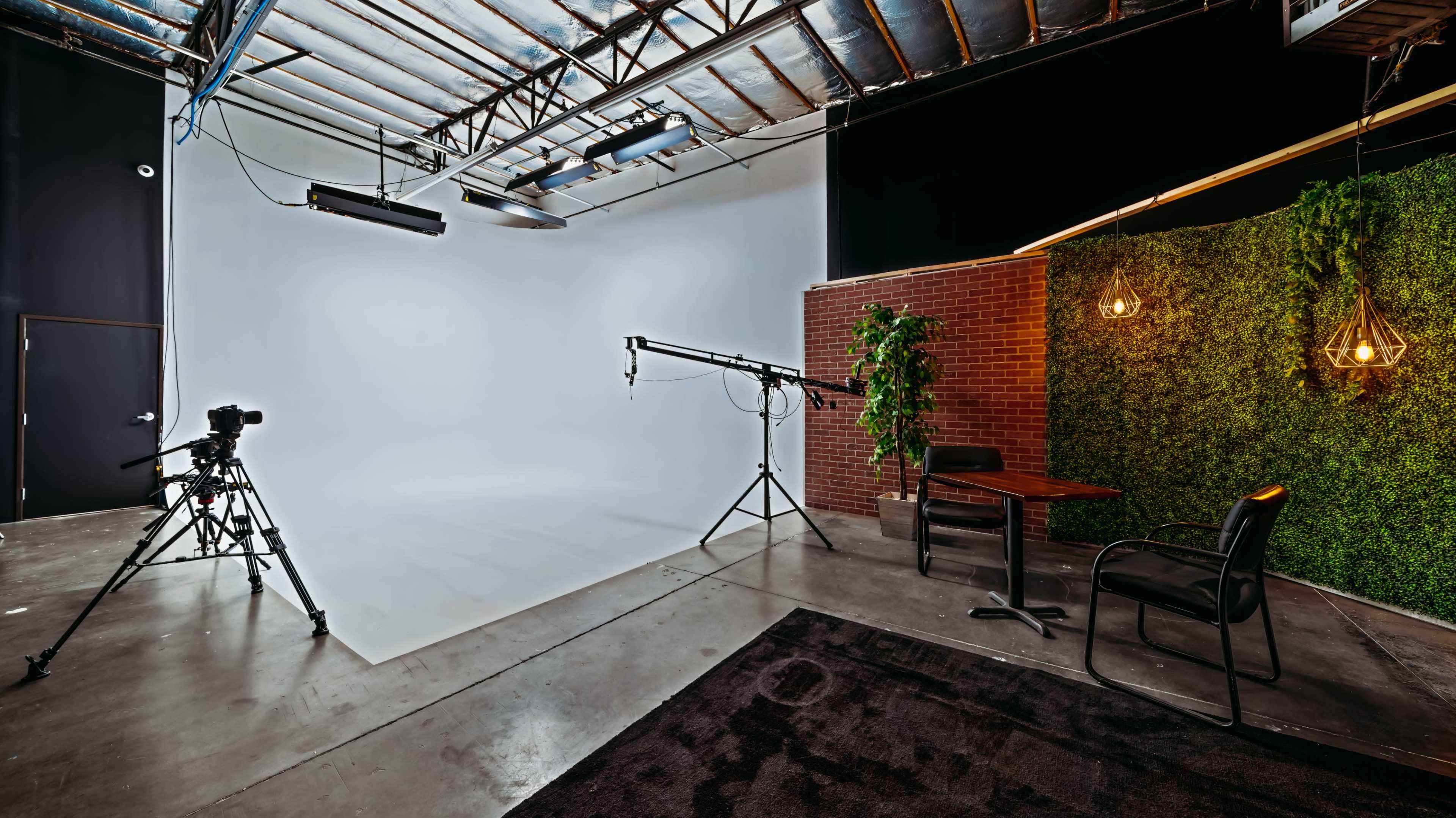 A photography studio with a plain backdrop, camera equipment, a table and chair, and a green plant wall.