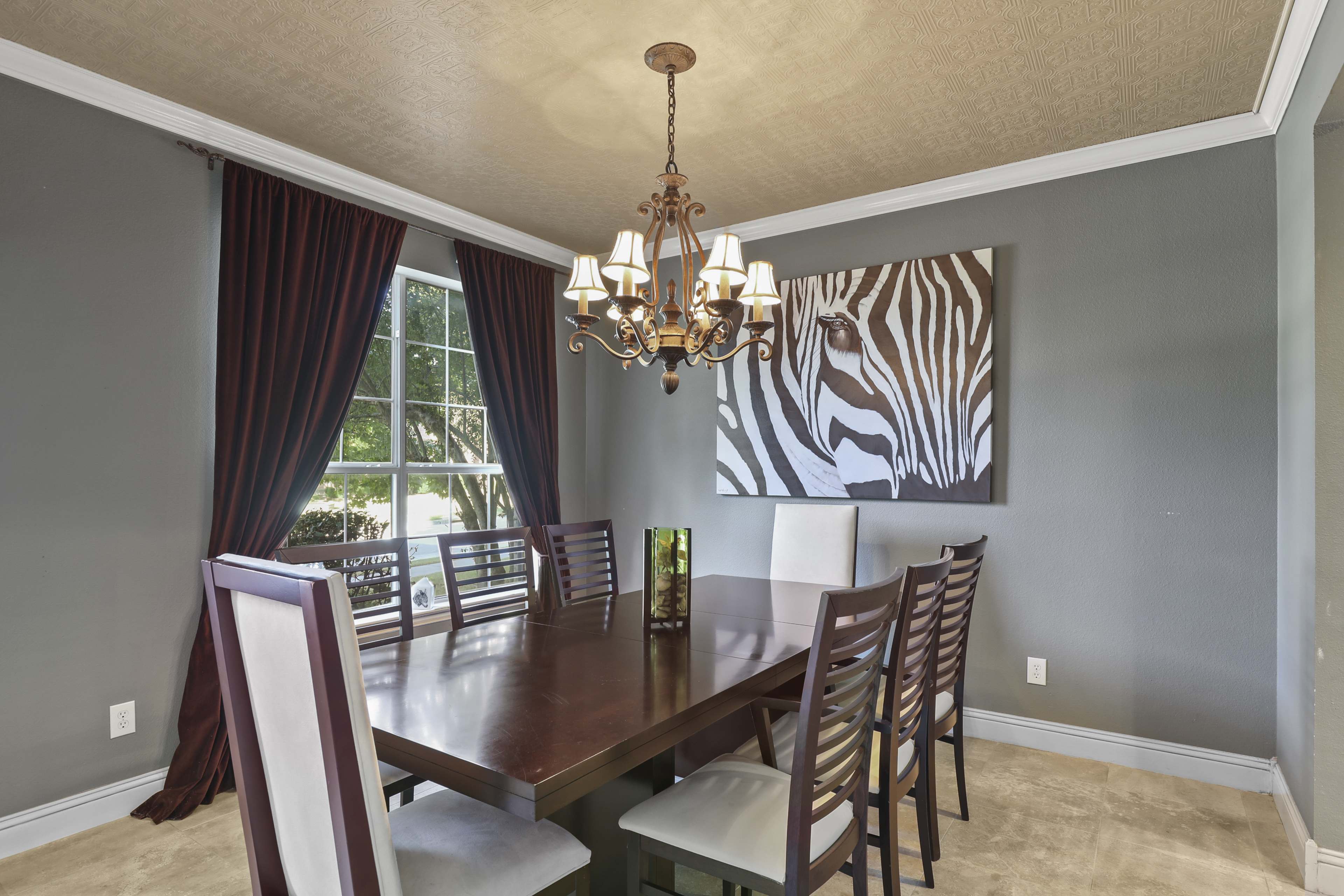 A dining room features a large wooden table surrounded by eight chairs, with a chandelier overhead and a zebra-striped artwork on the wall.