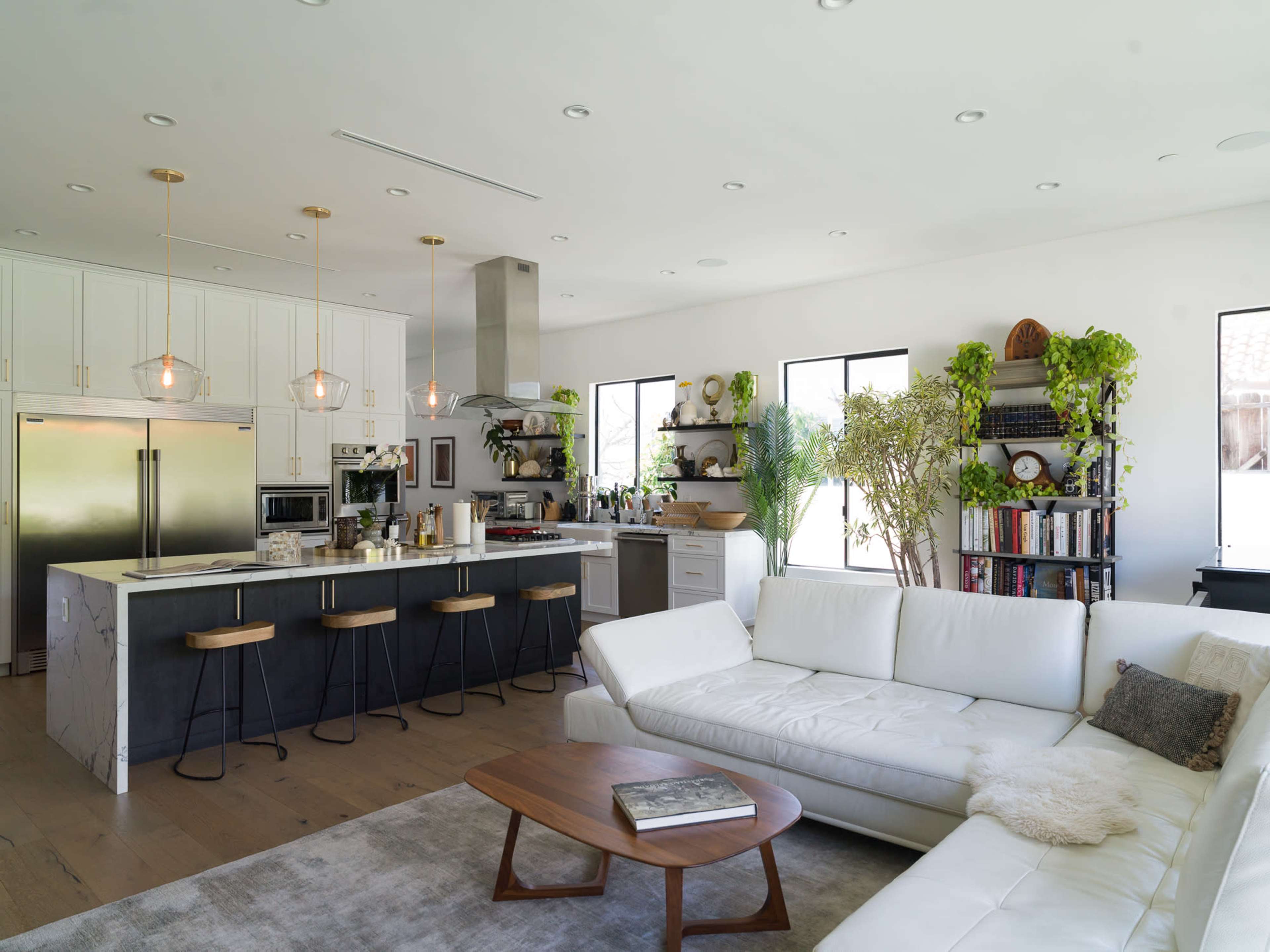 The image shows a modern kitchen and living room area featuring a white sectional sofa, a wooden coffee table, and a kitchen with stainless steel appliances and bar seating.