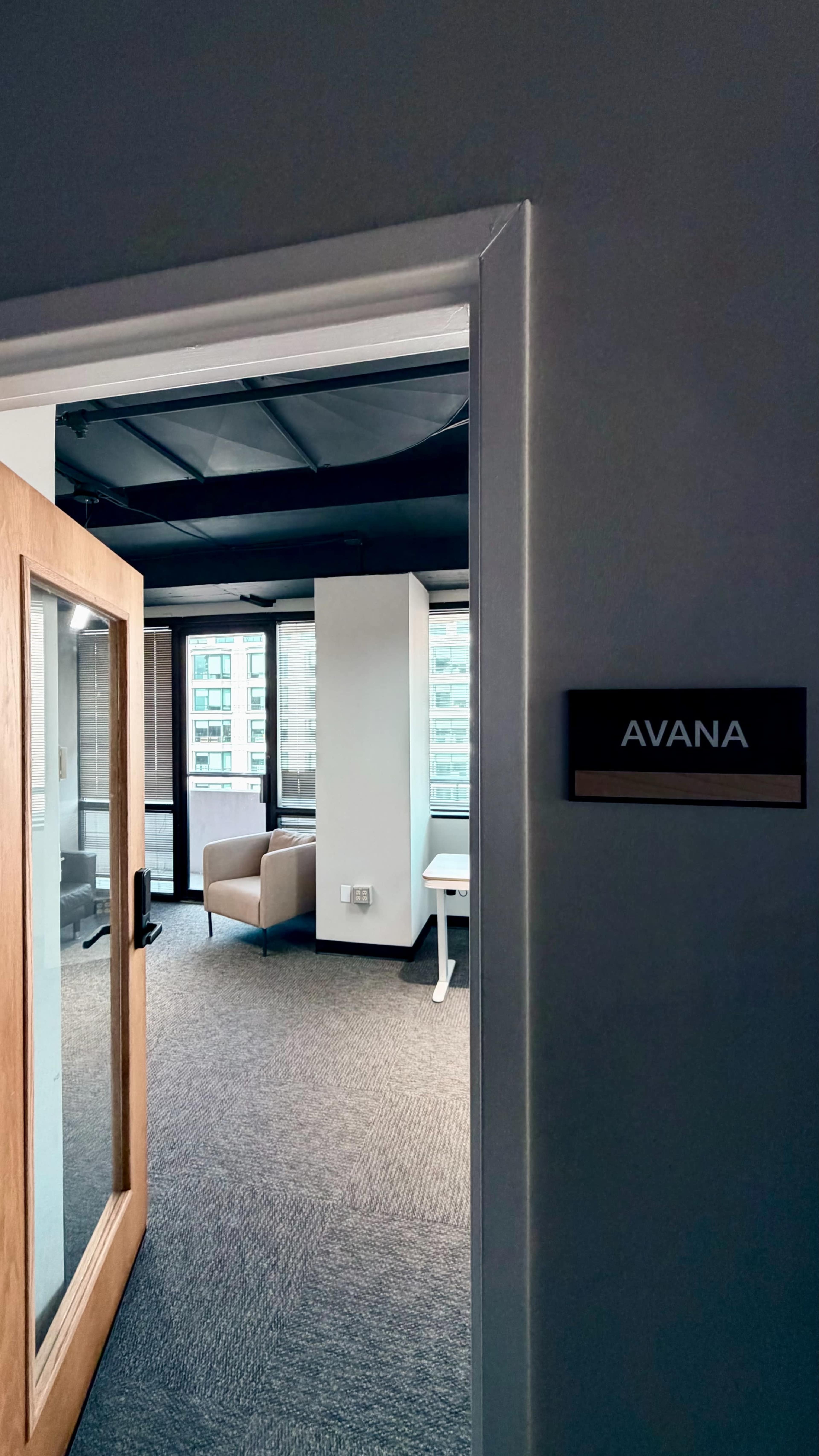 The image shows an office entrance with a sign labeled "AVANA," leading into a space that contains two armchairs and a table, visible through a glass-paneled door.
