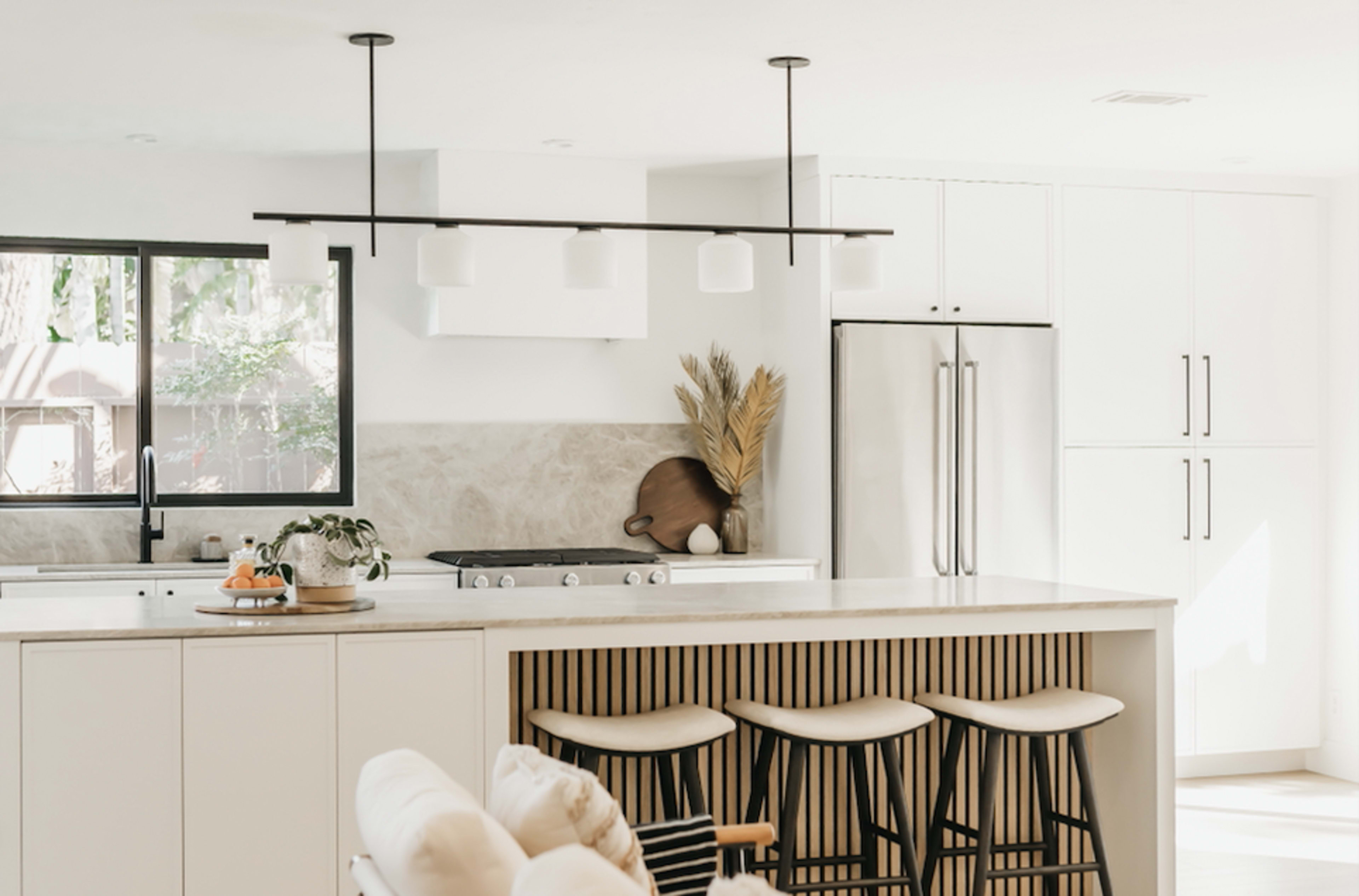 A modern kitchen features a central island with bar stools, white cabinetry, and a large window allowing natural light to illuminate the space.