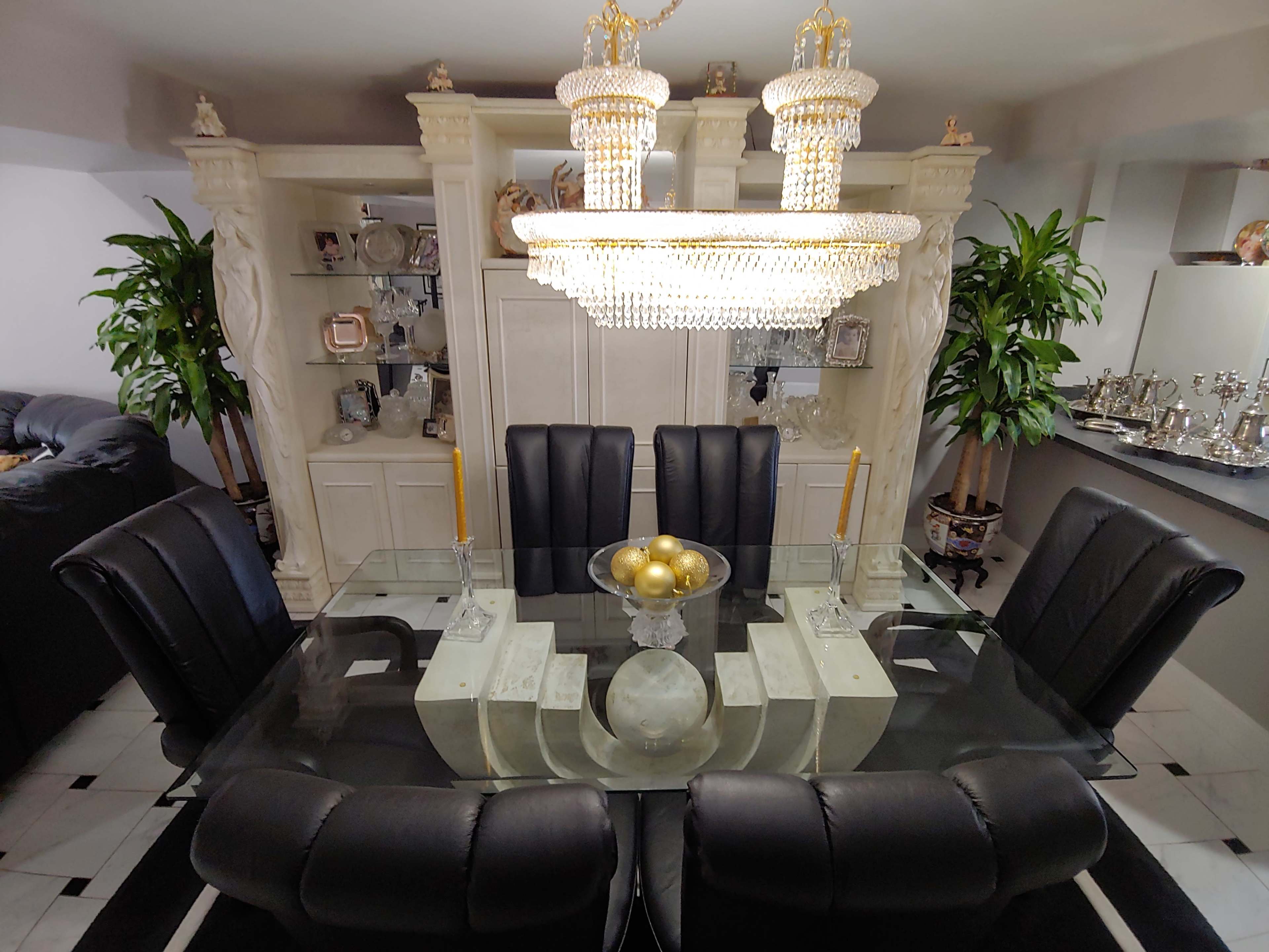 A glass dining table surrounded by black leather chairs, with a decorative bowl of gold ornaments and an elaborate chandelier overhead, situated near a white cabinetry backdrop adorned with decor.