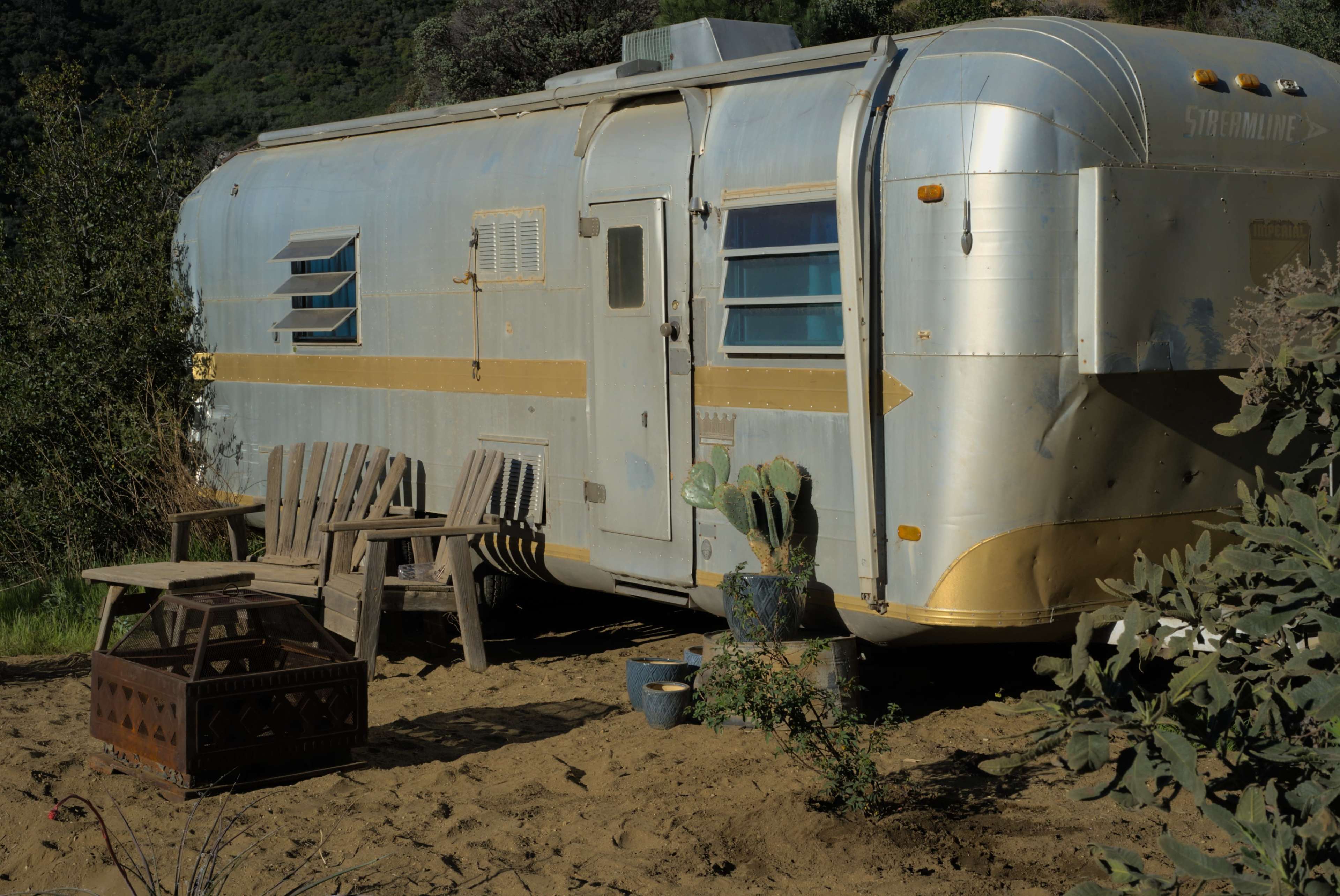 A vintage silver travel trailer is parked on sandy ground surrounded by desert vegetation, with two wooden chairs and a small table nearby.