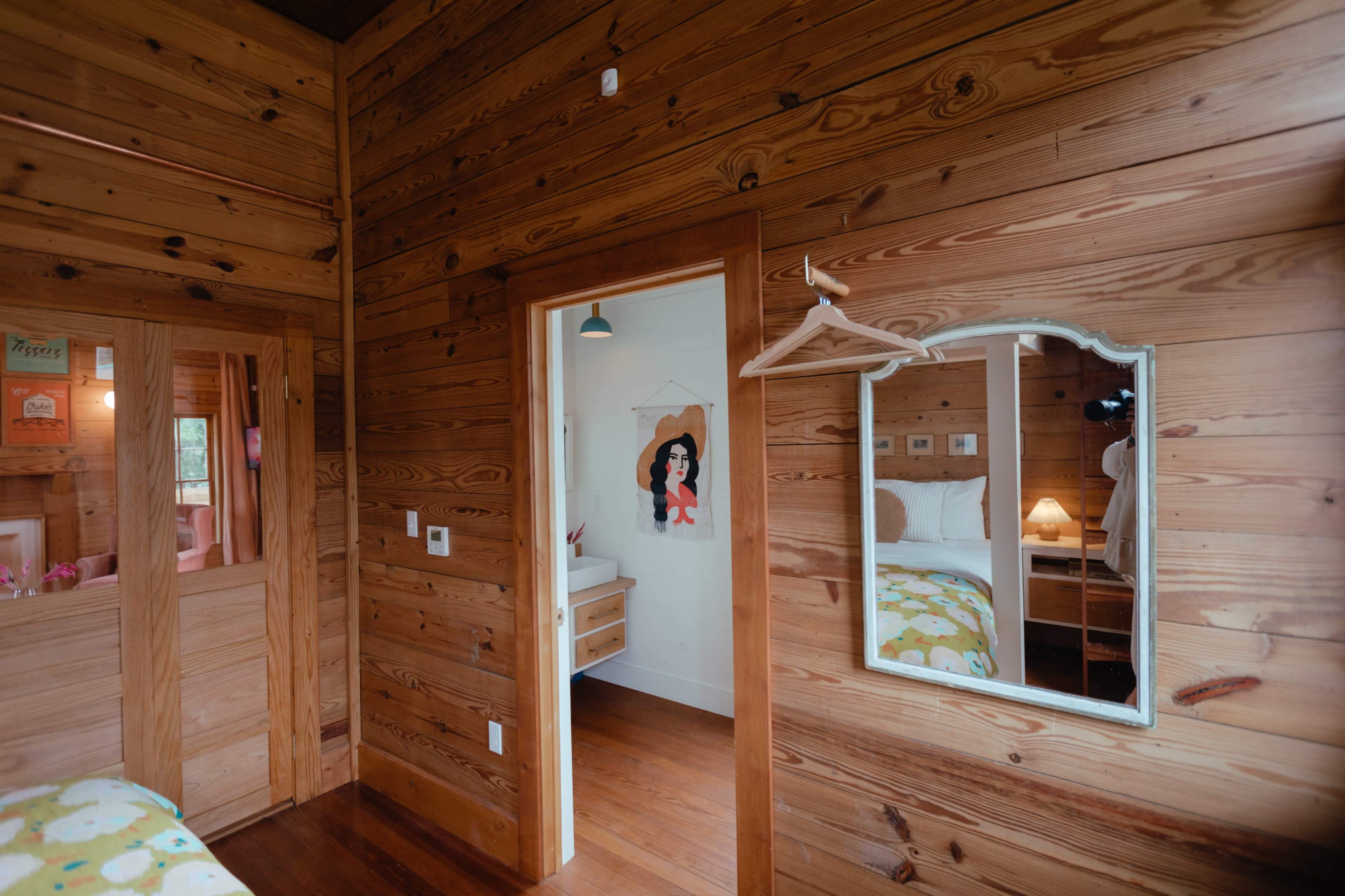 A wooden interior of a small room featuring a mirror, a door leading to another space, and a wall adorned with a poster and a coat hook.