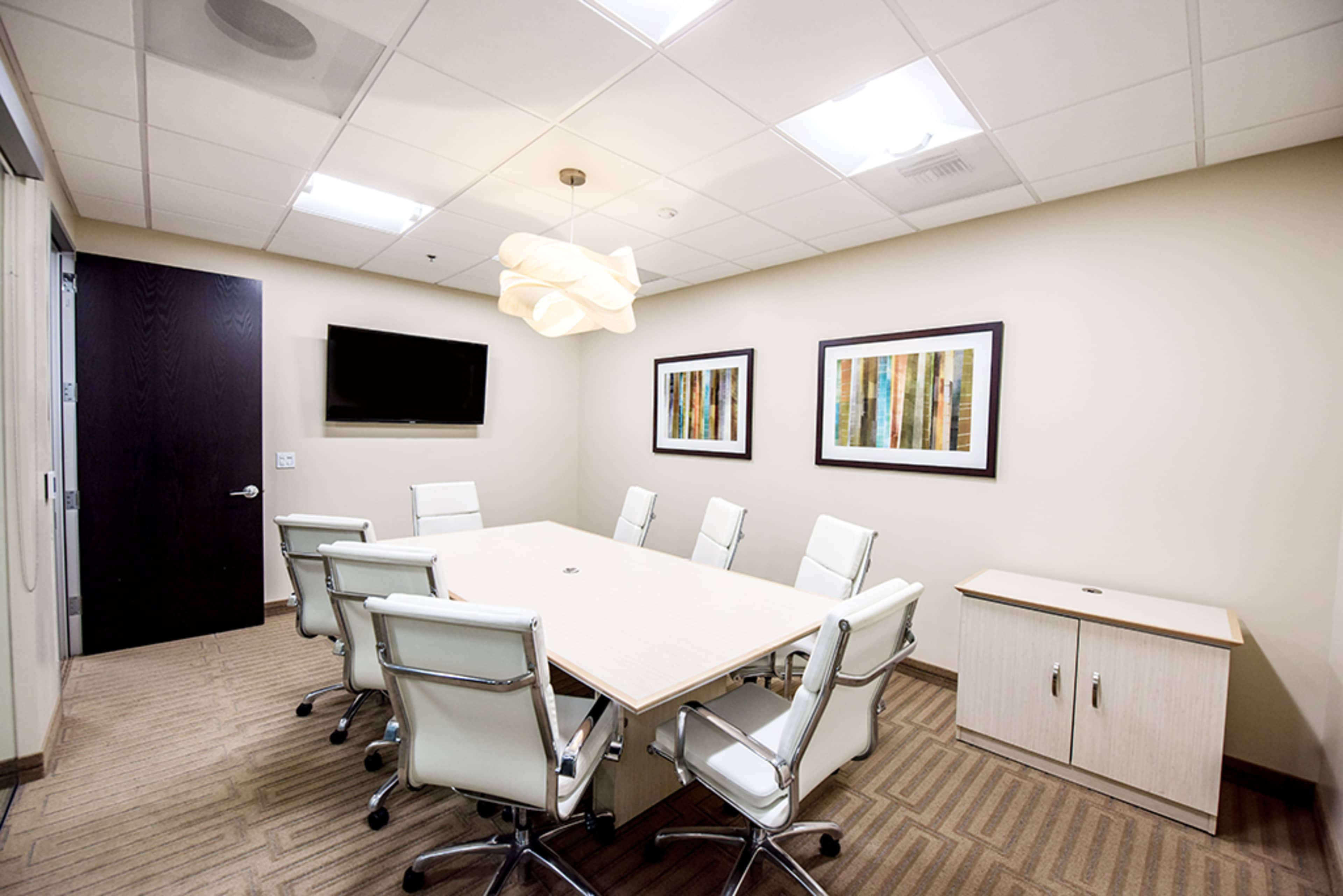 A modern conference room features a large rectangular table surrounded by white leather chairs, a mounted television, and two framed artworks on the light-colored walls.