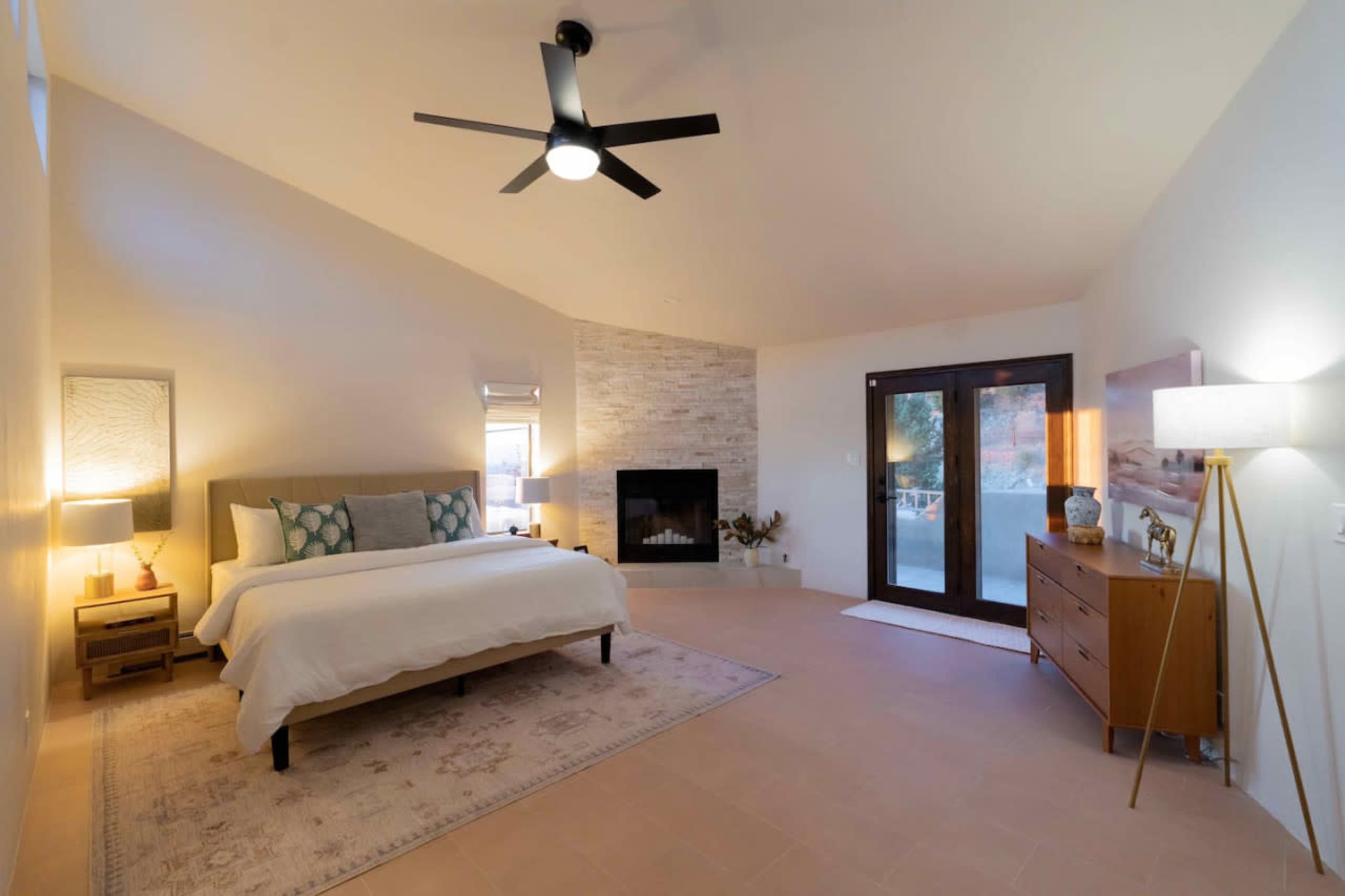 The room features a bed with decorative pillows, a minimalist design, a fireplace, double doors leading outside, and a dresser with a lamp.