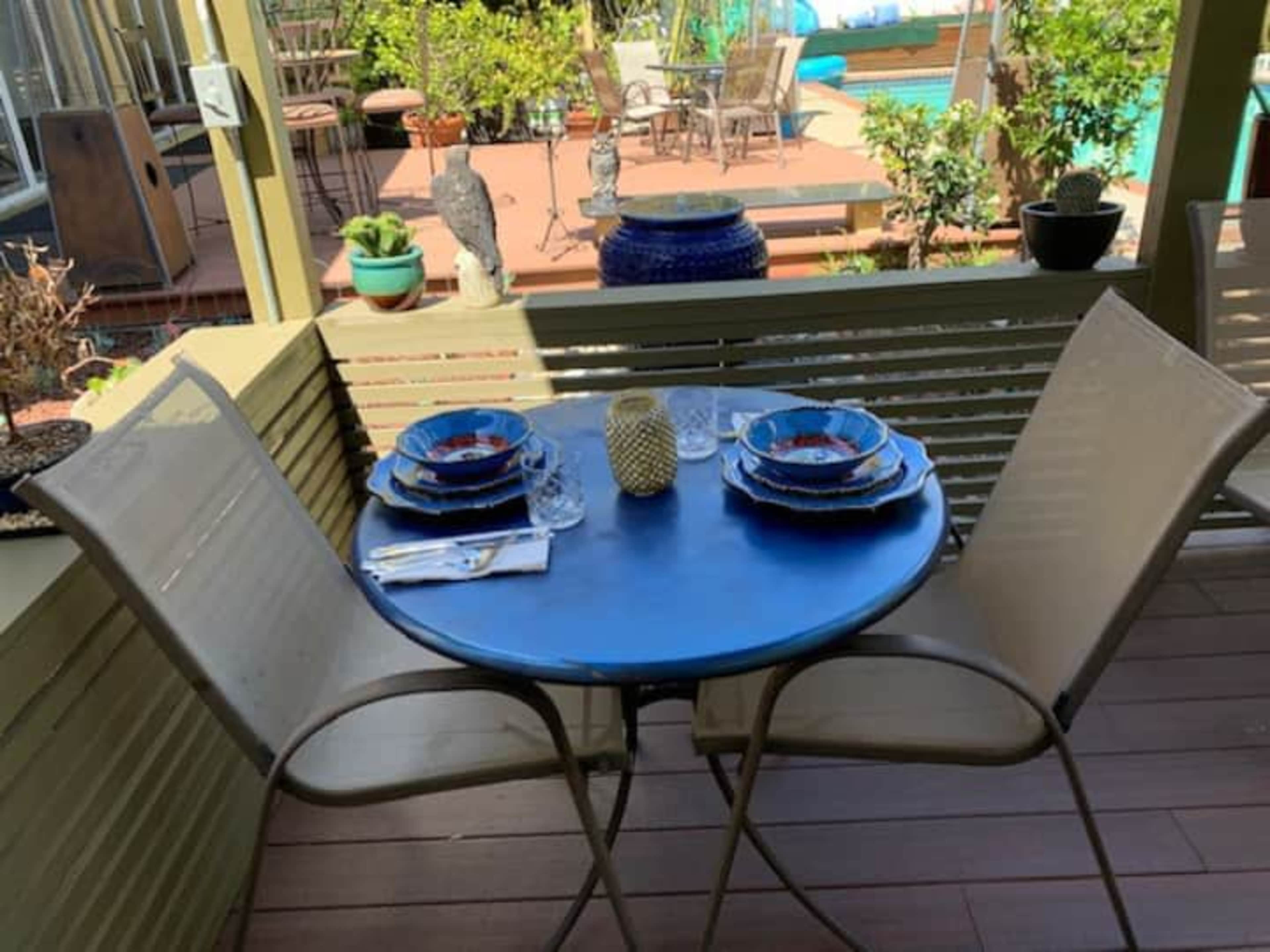 A round blue table with two chairs is set for a meal with plates and glasses, surrounded by greenery and a view of a pool in the background.