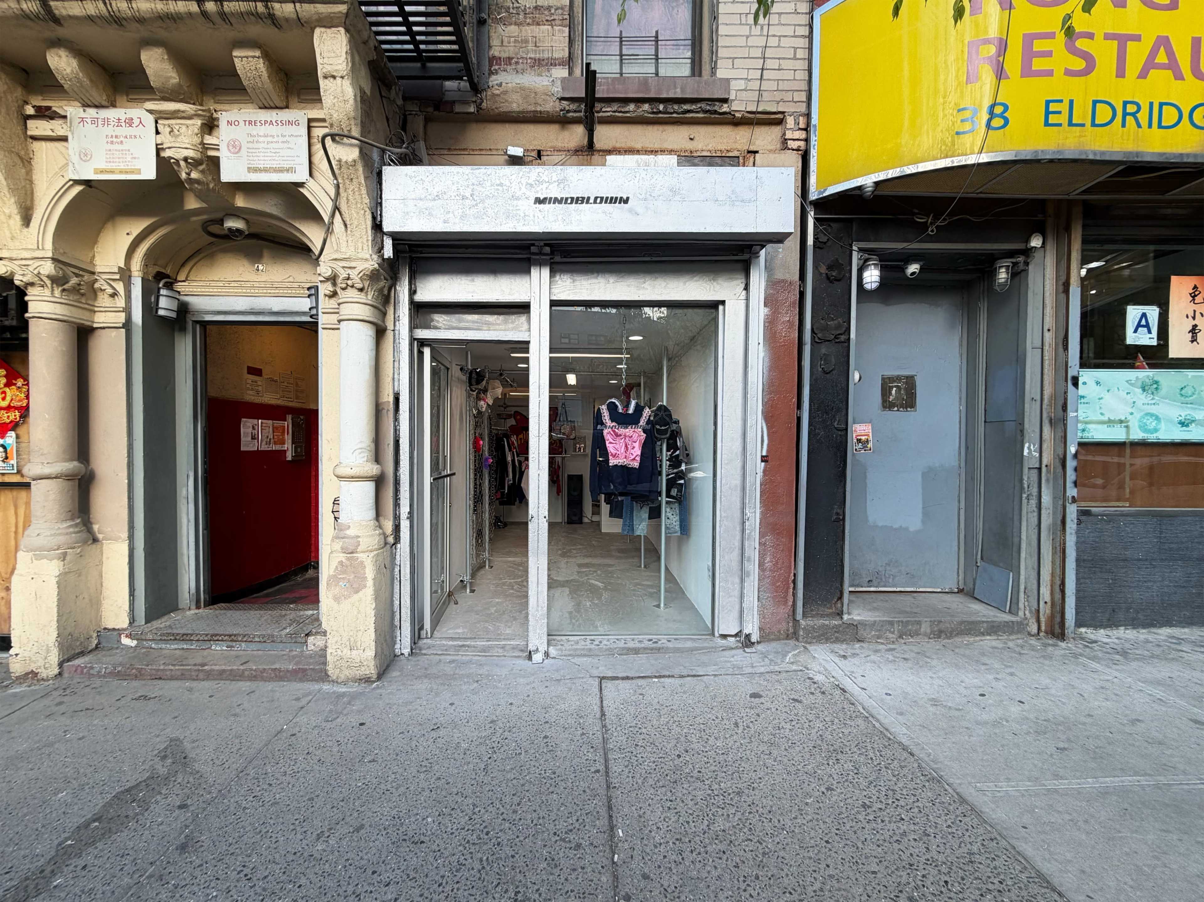 The image shows a retail storefront with large glass doors and a display of clothing visible inside, situated next to a restaurant entrance on a city street.