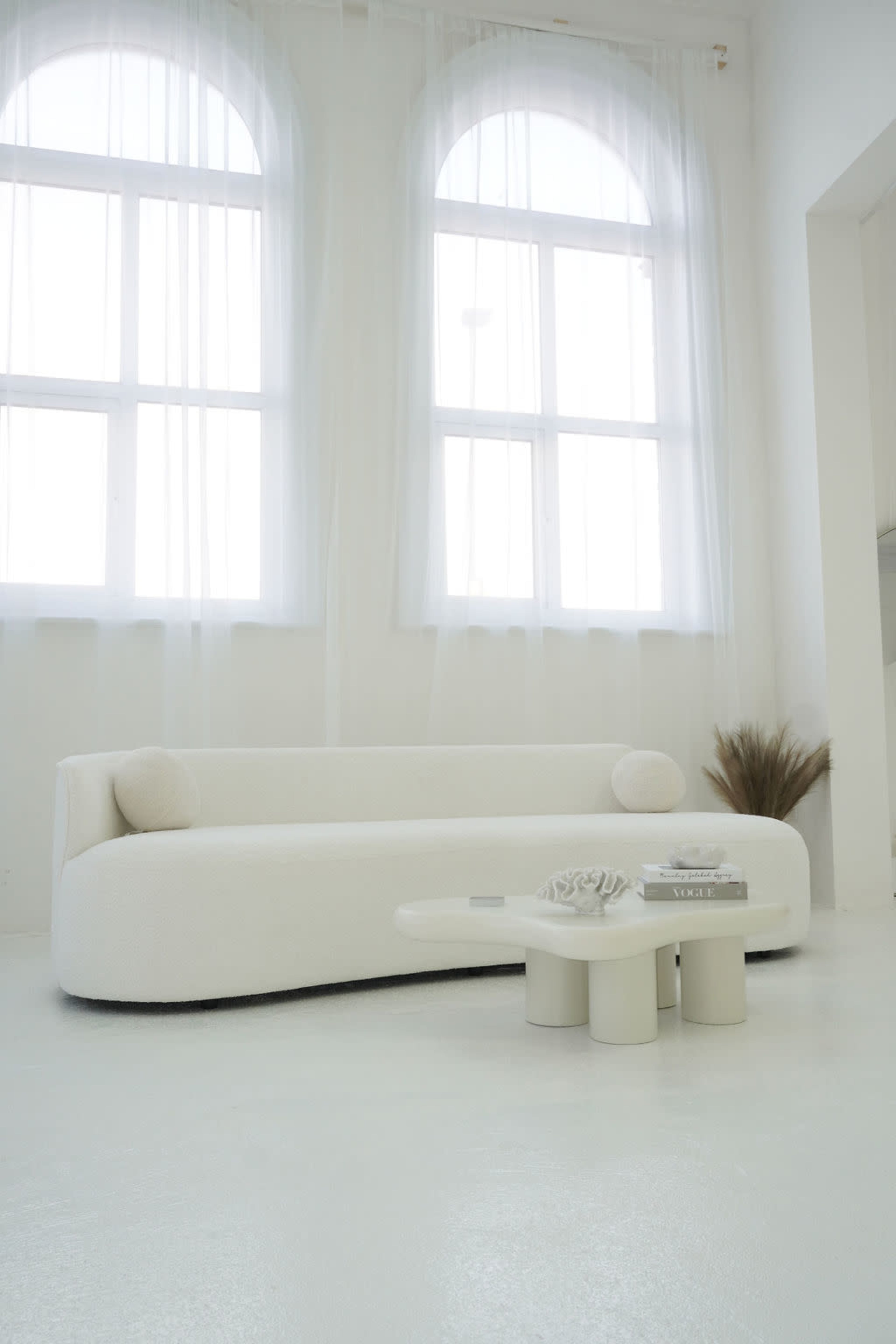 The image features a modern, white, curved sofa positioned in a bright room with large arched windows and minimal decor.