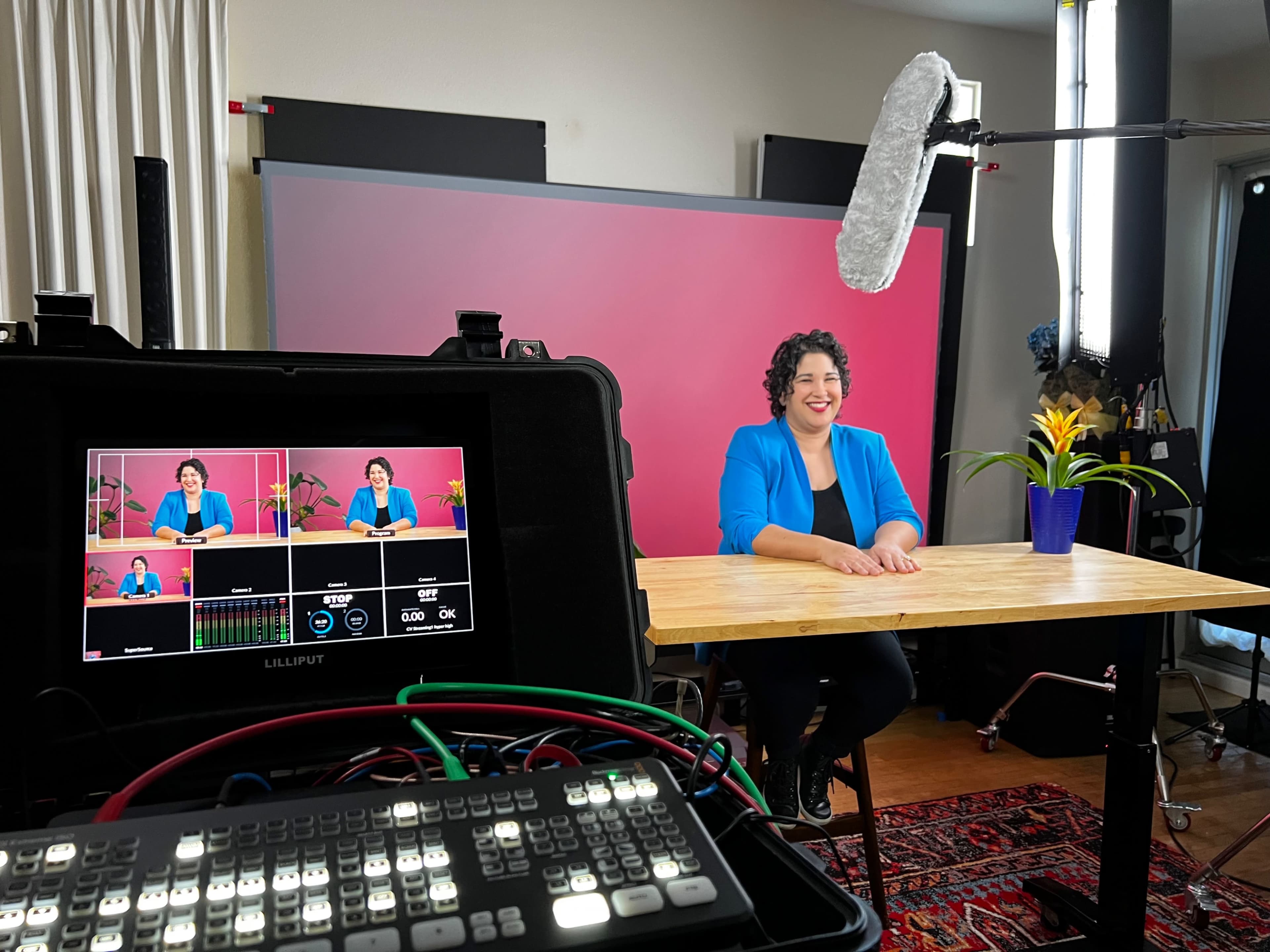 A woman in a blue blazer sits at a wooden table in a studio setup with a pink backdrop and a microphone overhead.