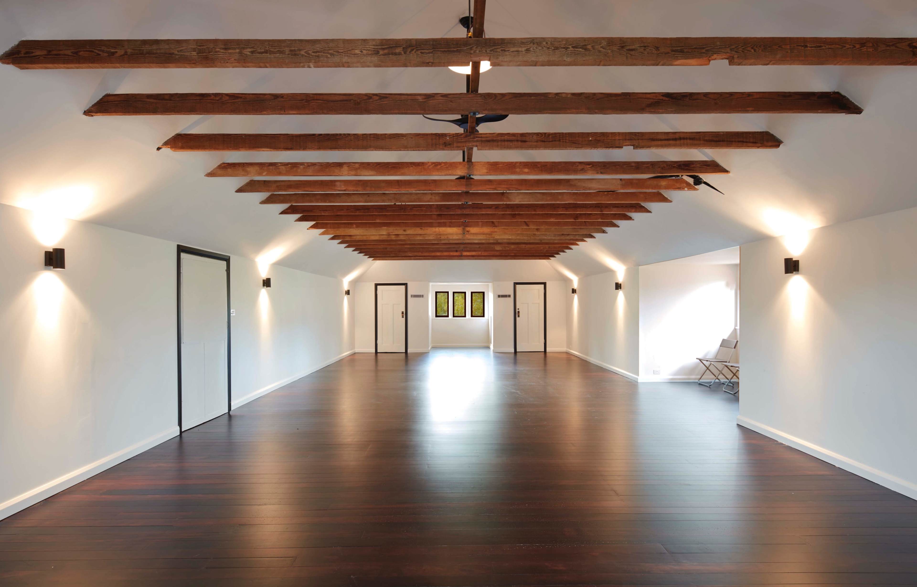 A spacious, well-lit interior room with wooden beams on the ceiling and dark hardwood flooring, featuring several doors on one side and art displayed on the opposite wall.