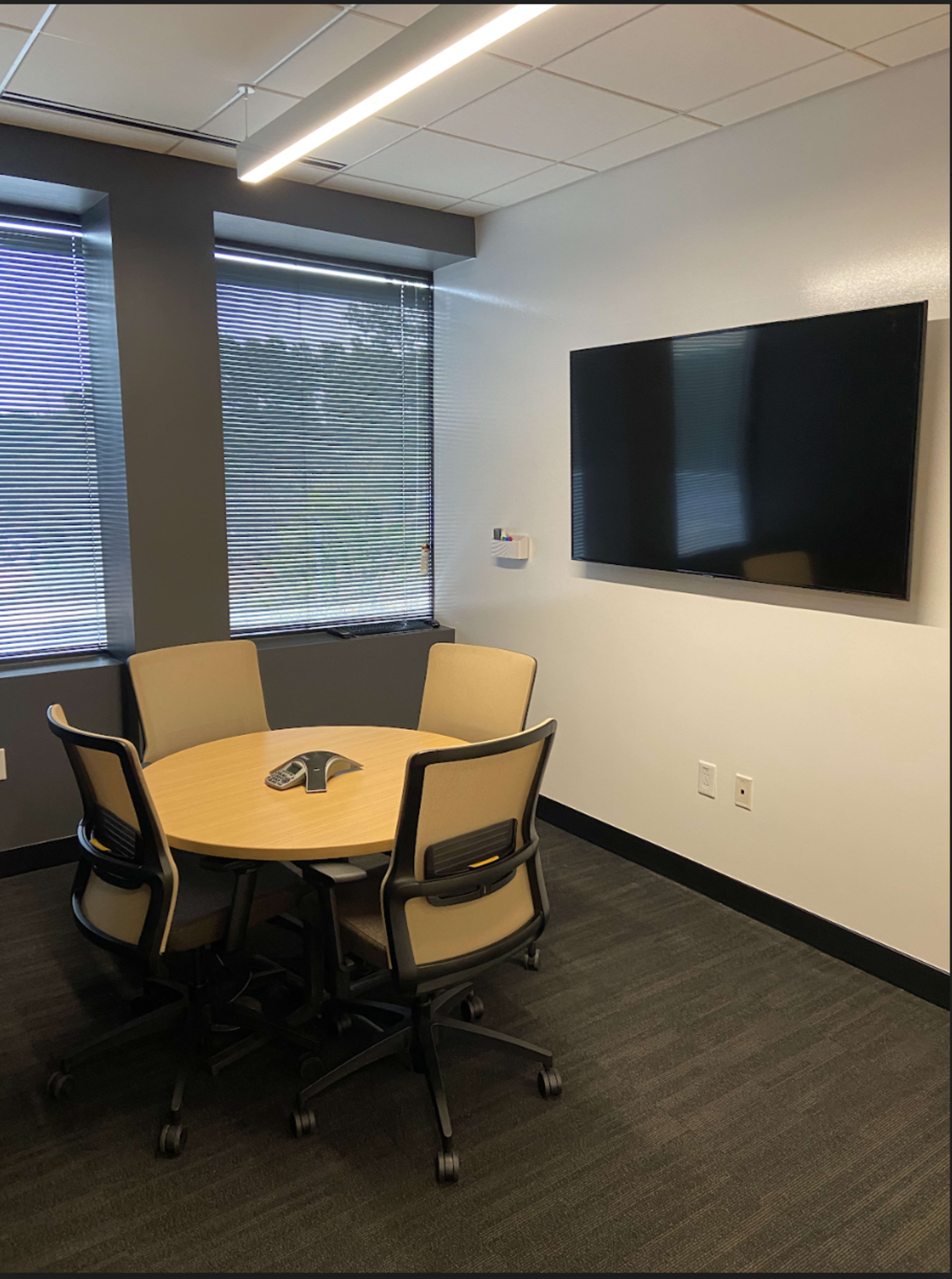 A small conference room features a round table with four chairs, a wall-mounted TV, and windows with blinds.
