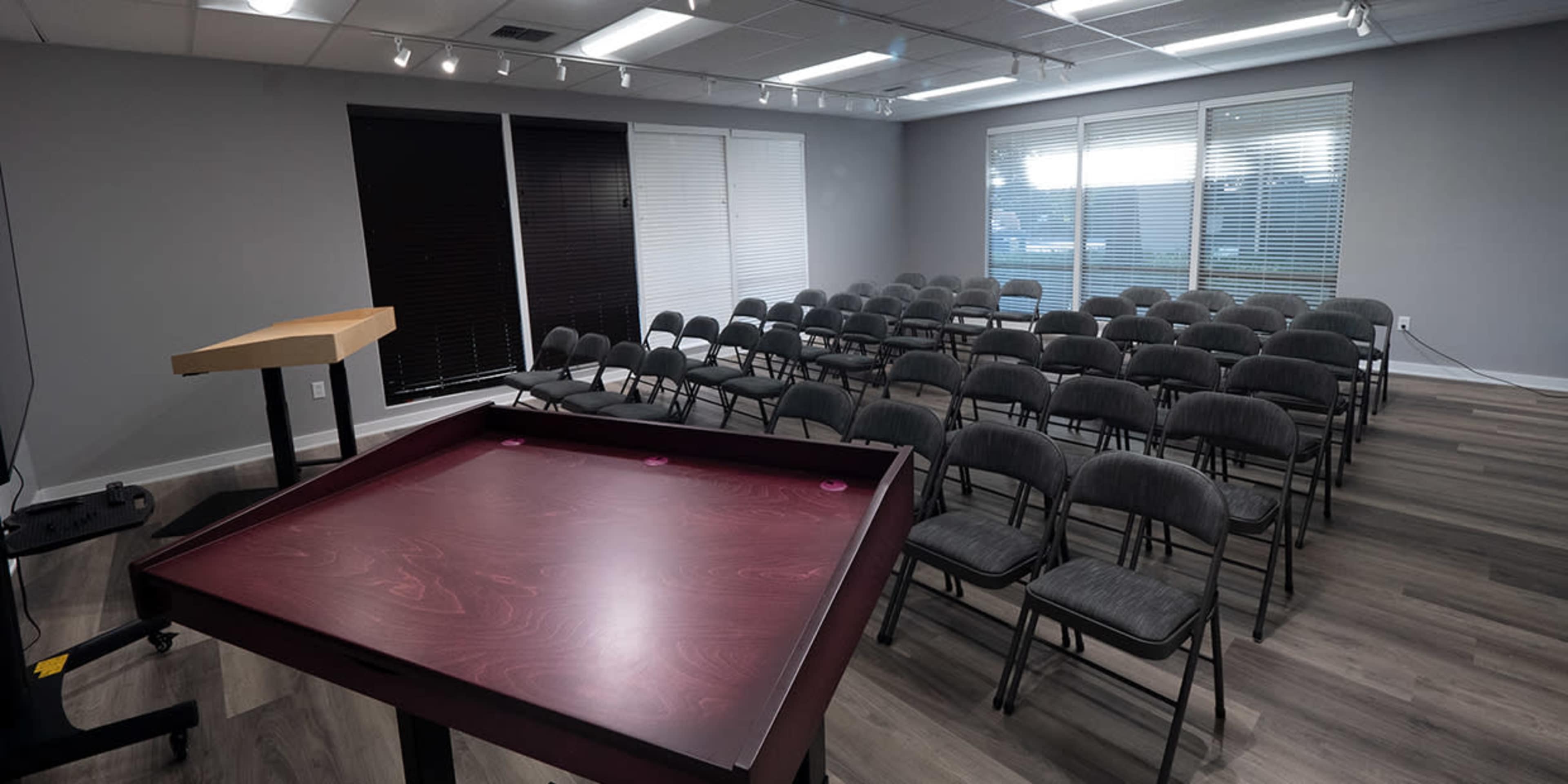 A conference room features a wooden presentation table in the foreground and rows of gray folding chairs arranged for an audience.