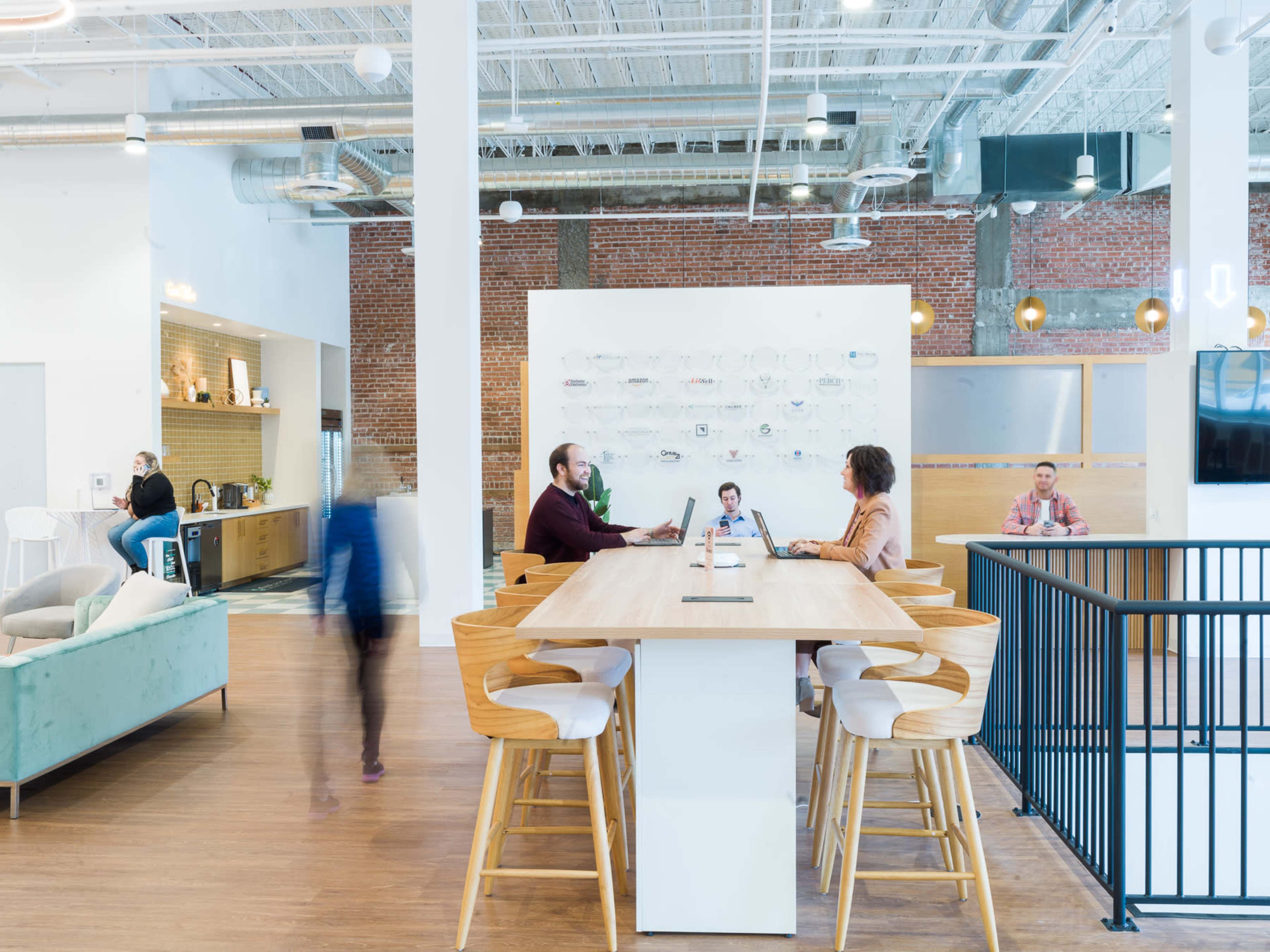 The image shows a modern office space with people working at a long table, a lounge area, and exposed brick walls.