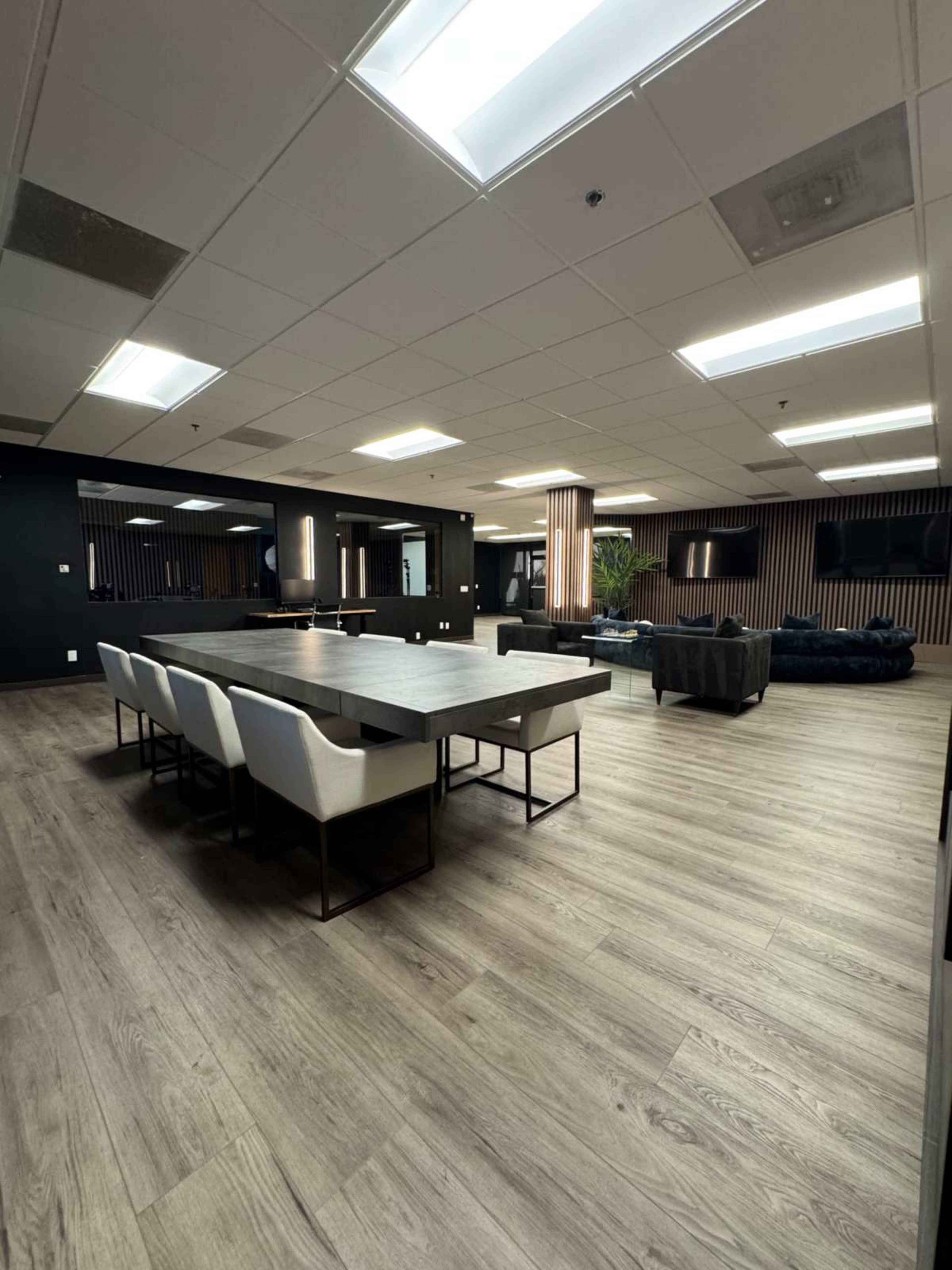The image shows a spacious, modern office interior featuring a large dining table with white chairs, a seating area with a dark sofa, and wooden flooring.