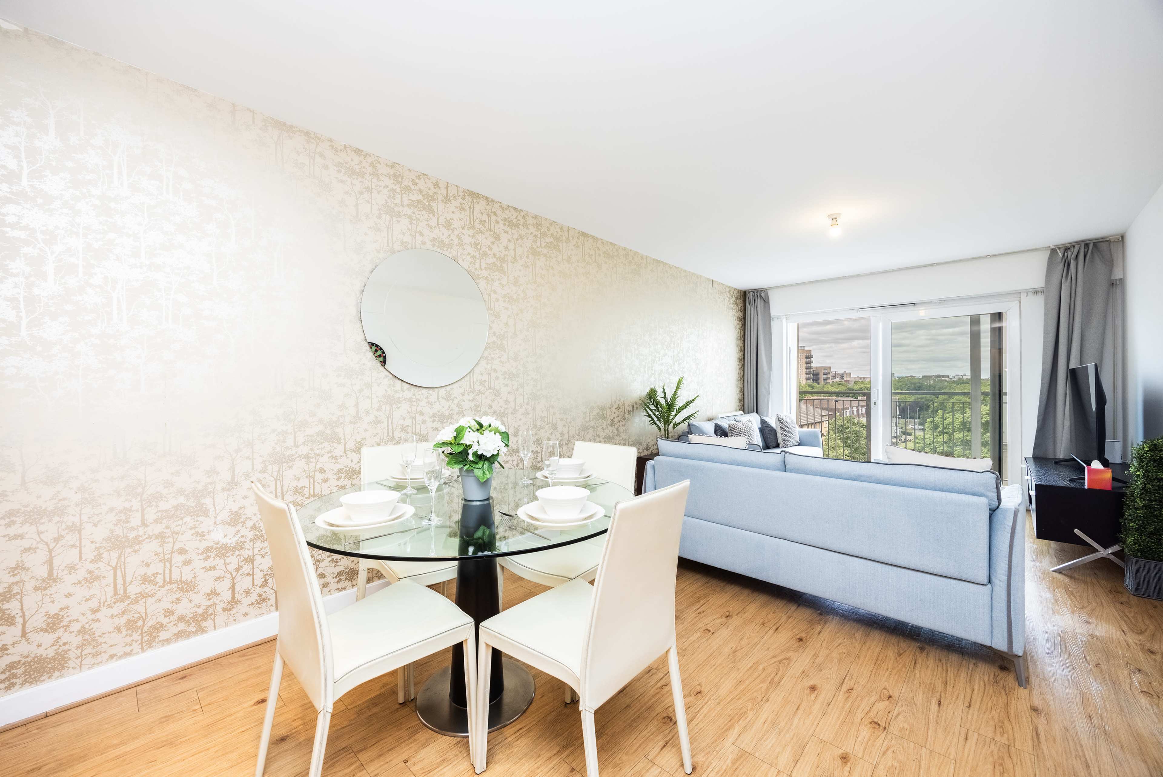 A dining area with a glass table and four white chairs is positioned next to a living area featuring a gray sofa and large windows overlooking a green landscape.