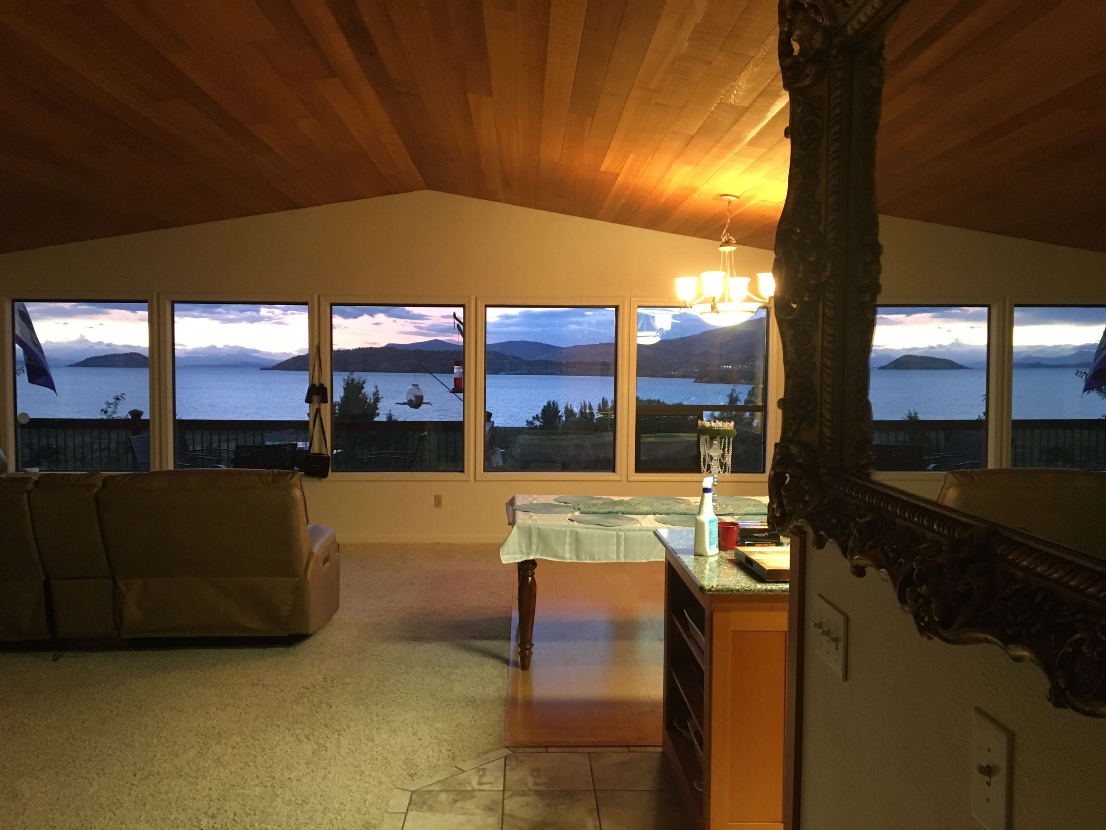 The interior of a home features a kitchen and living area with large windows displaying a view of a lake and mountains at sunset.