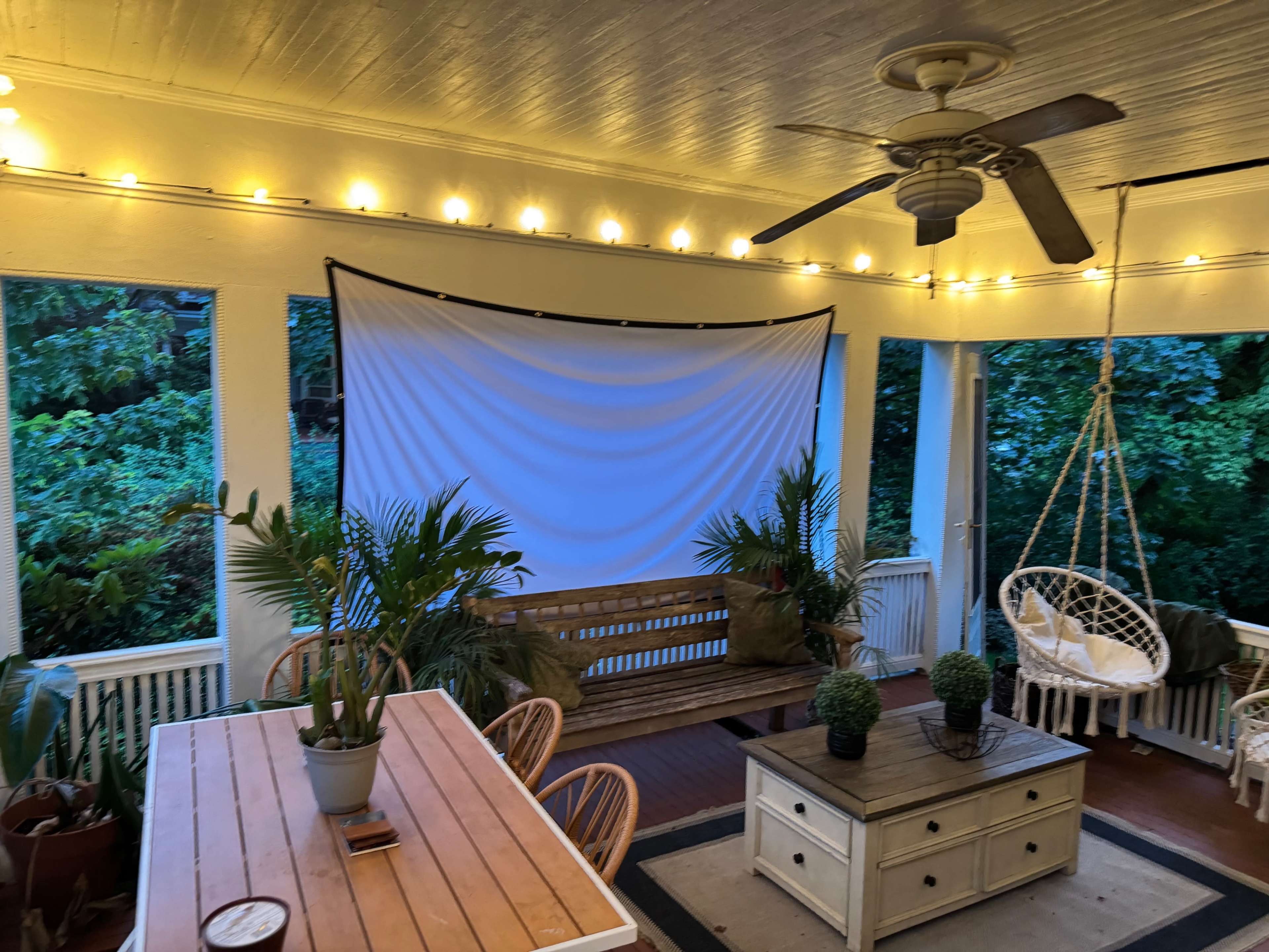 The image shows a cozy outdoor porch with string lights, a large screen, a wooden bench, a hanging chair, and potted plants.