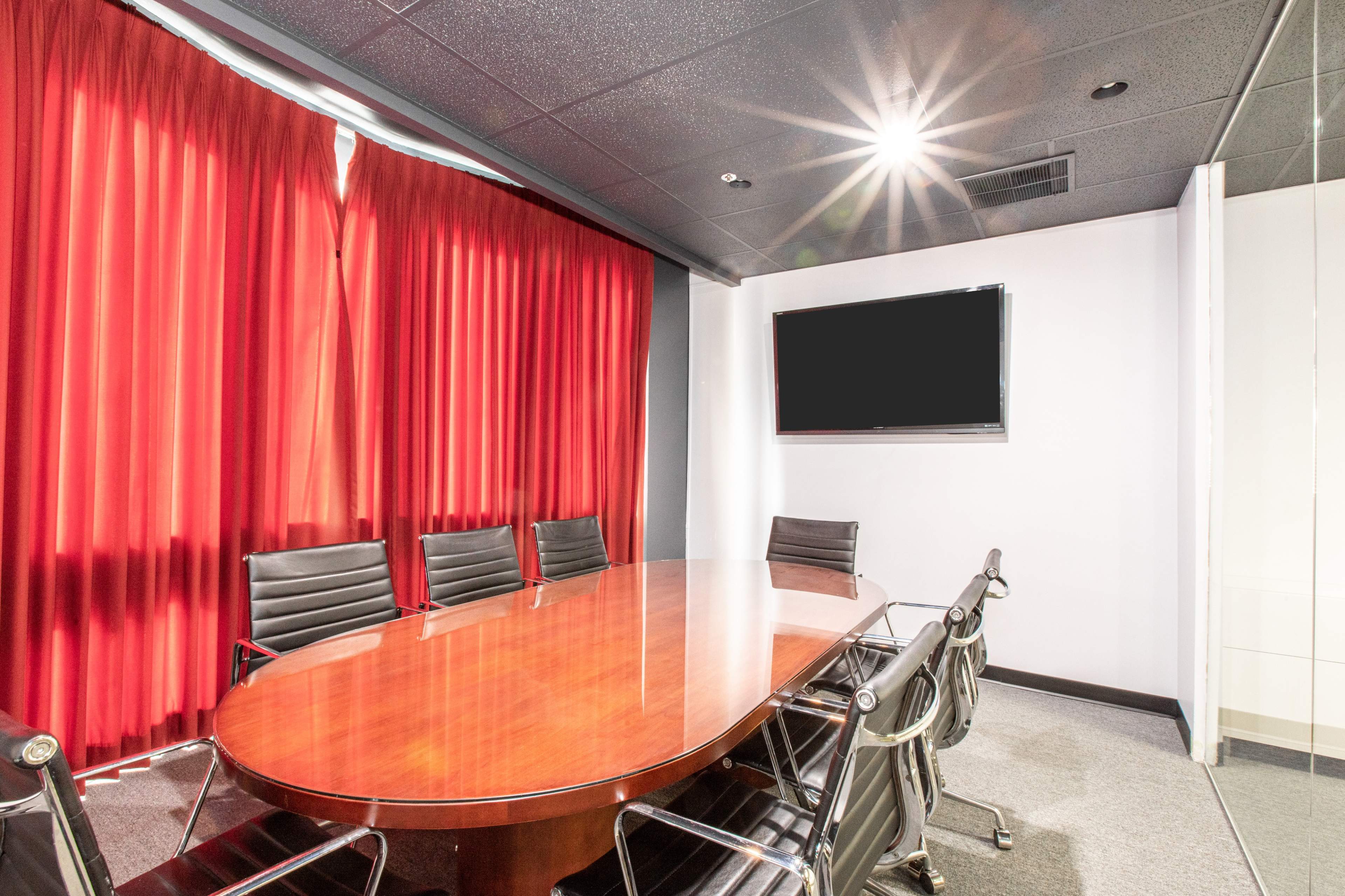 The image shows a conference room with a large oval wooden table surrounded by black leather chairs, red curtains in the background, and a wall-mounted TV.