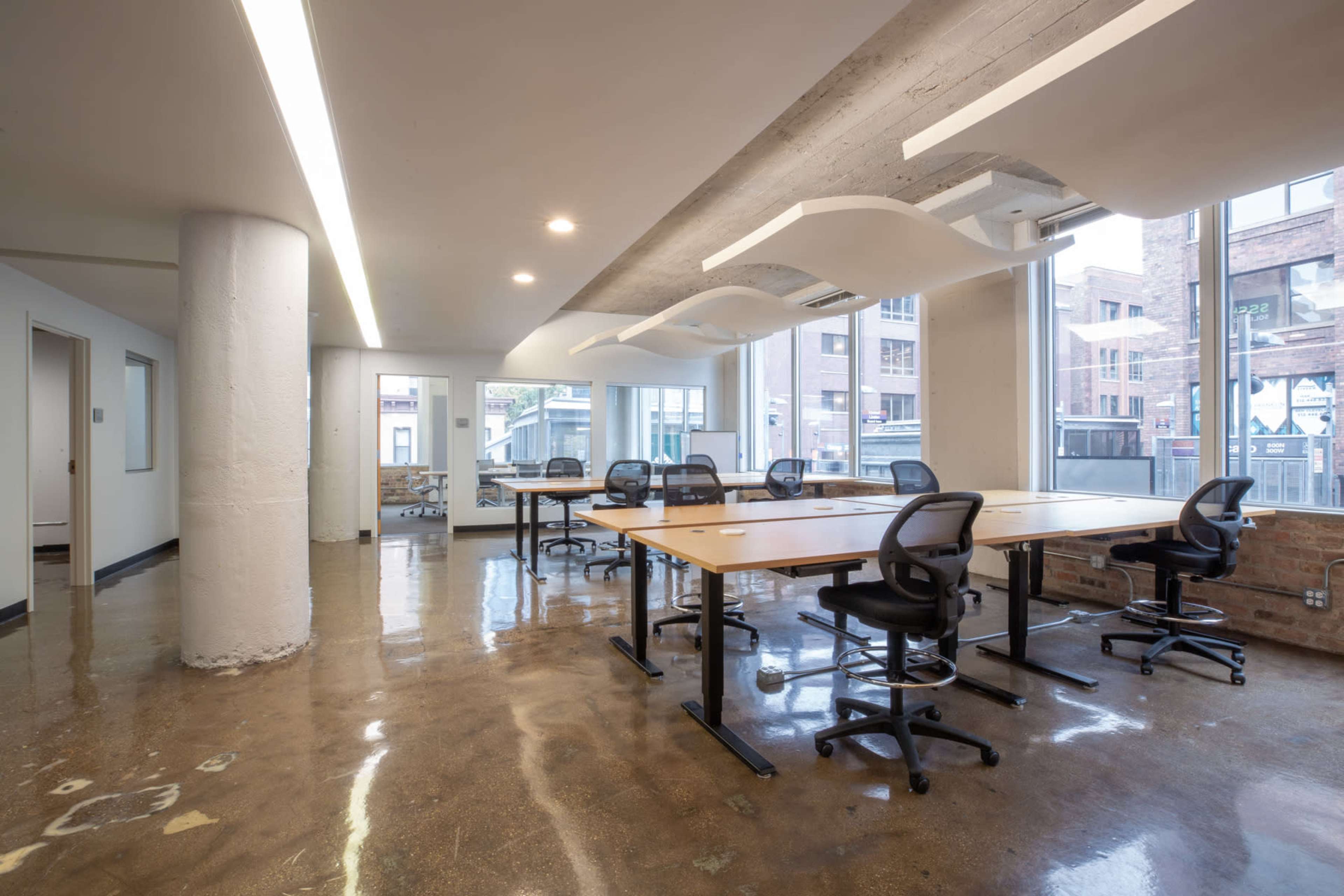 An open office space features several desks and chairs arranged neatly, with large windows allowing natural light to enter.